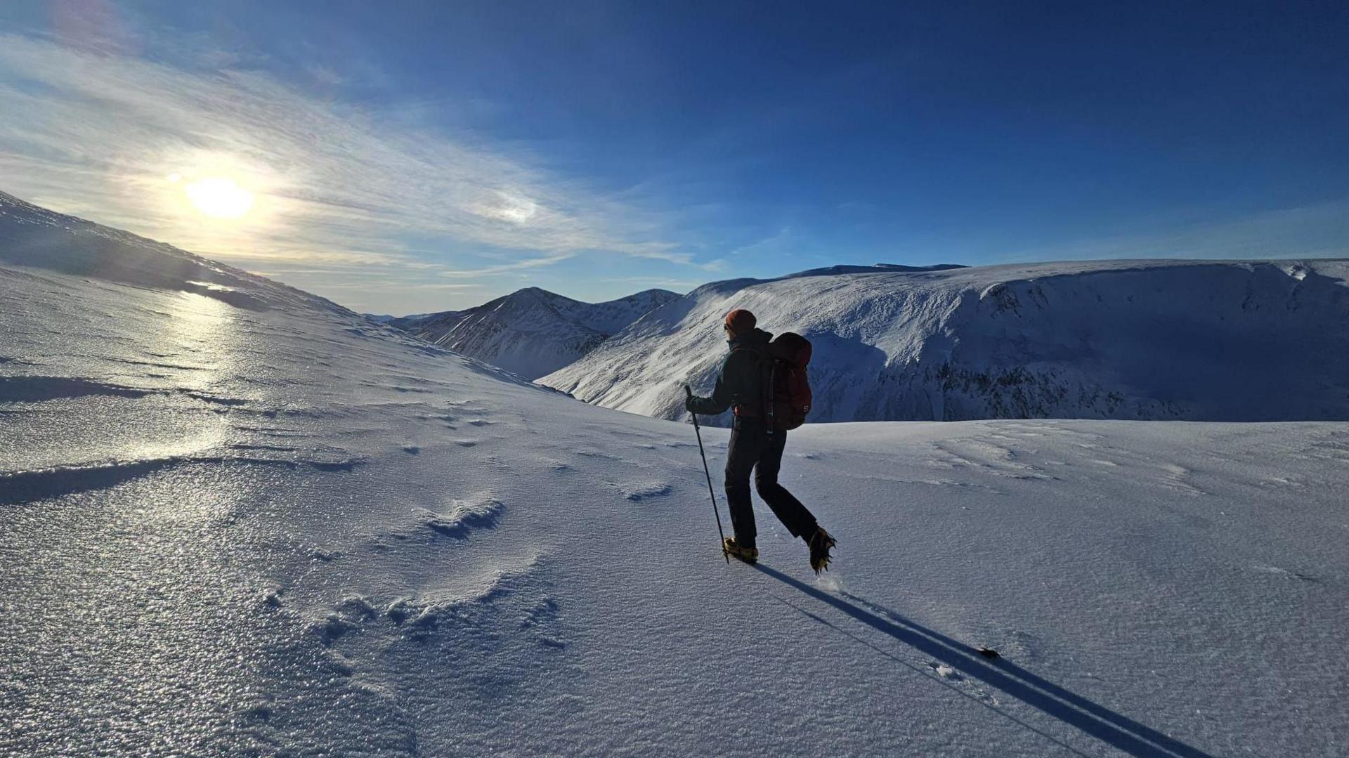 A single walker crosses the snow and ice on the way to the top of the mountain, Ben Macdui in the Scottish Highlands. They are walking towards the sunshine and the whole countryside is covered in white snow.