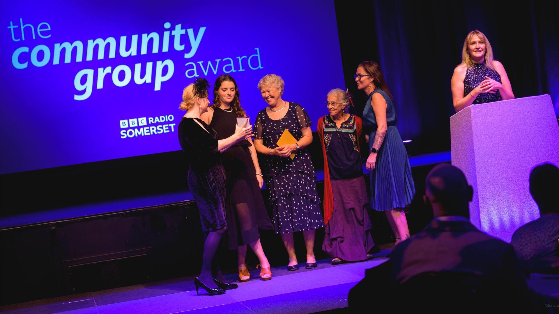 Lady shows four other women on stage the trophy they have won to the left of presenter who is standing behind podium