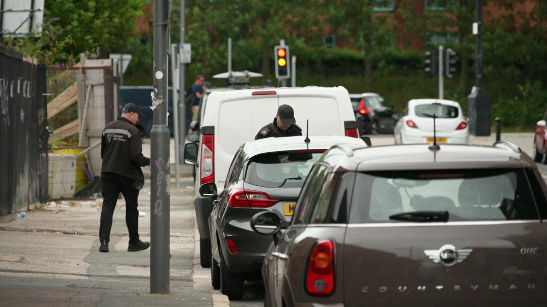 Cars are parked in on-street bays. Traffic wardens are checking on vehicles.