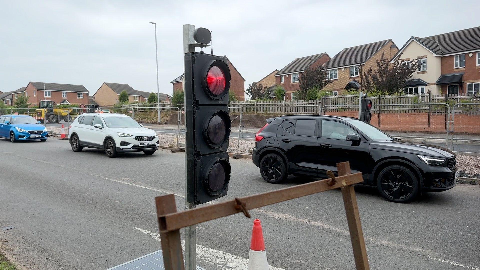 A temporary traffic light on red as cars drive past in the background. Behind the cars are metal fences blocking off a roadworks site. A row of houses is visible behind a wall on the other side of the road.