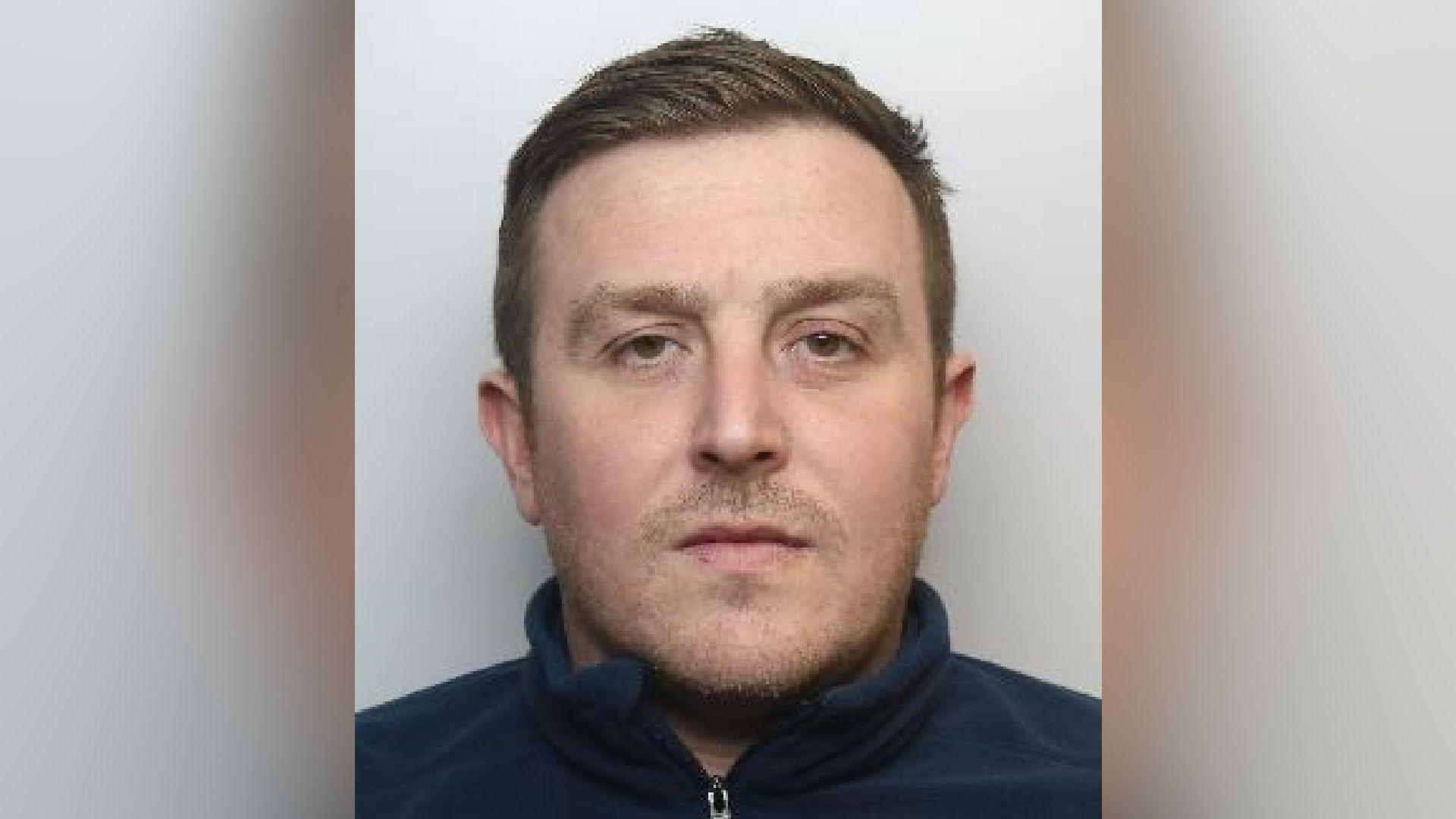 A mugshot of a man who has his brown hair combed over to one side. He has facial hair and is wearing a blue sweater.