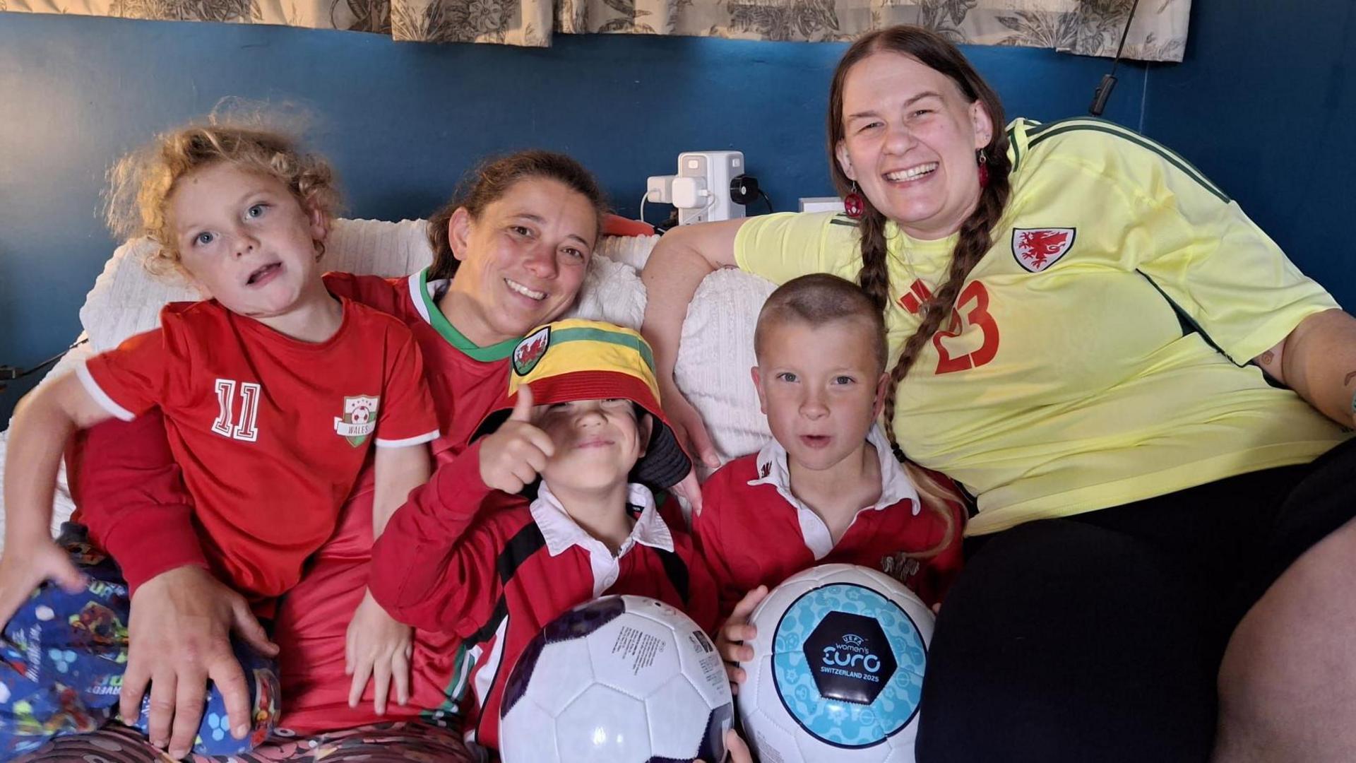 A woman is on the right of the image in a yellow Wales shirt, smiling at the camera and her hair is in pigtails. There are three boys all in red Wales shirts and the two sat closest to the woman have footballs in their hands. The one in the middle has a bucket hat on and his thumb up tot the camera. Next to him is a woman with a red Wales shirt and dark hair tied up smiling. She hold a boy on her lap with a red Wales shirt and blue pj bottoms who has blonde curly hair.