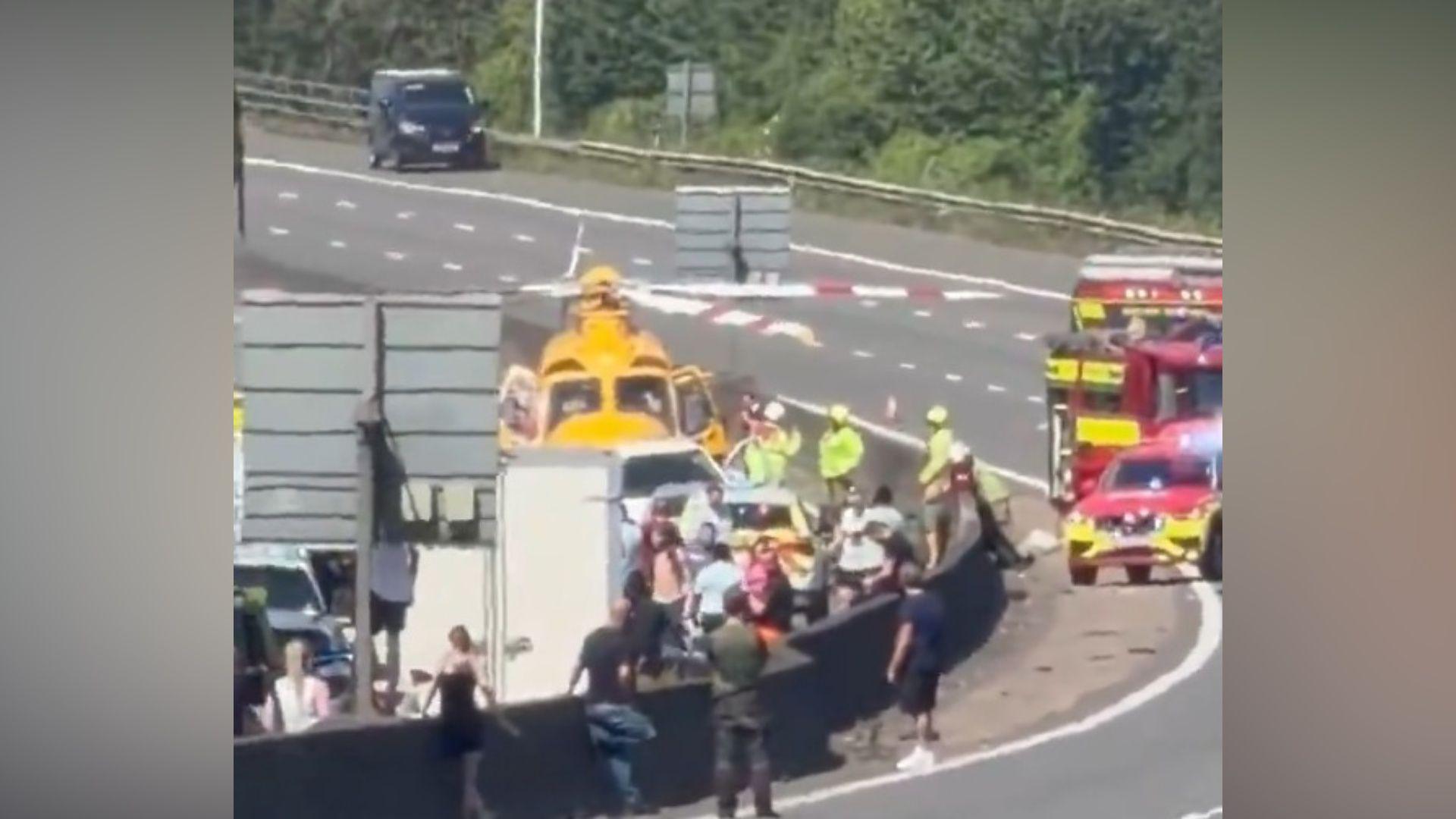 Emergency services and motorists spending on a motorway. There is a yellow helicopter nearby and two fire brigade vehicles.