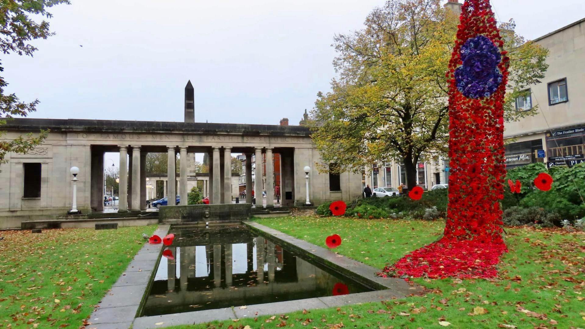 Red knitted poppies hang down in a drape, with purple knitted poppies forming one giant poppy within the drape. It hangs down in Remembrance gardens in Southport, on an autumn day