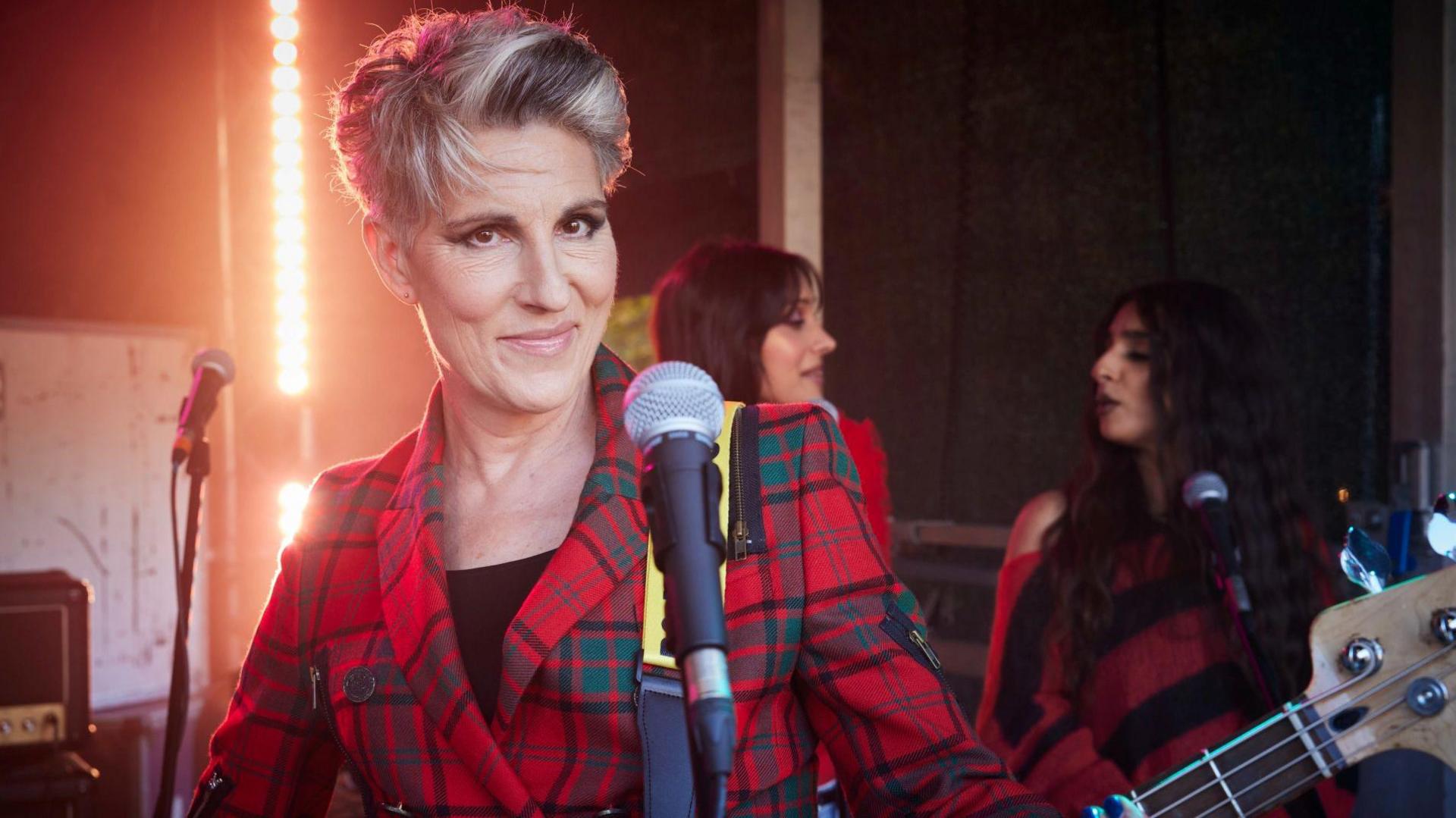 Tamsin Greig stands behind a microphone. She has grey hair, heavy eye make-up and is wearing a red tartan suit. She is holding a bass guitar.
