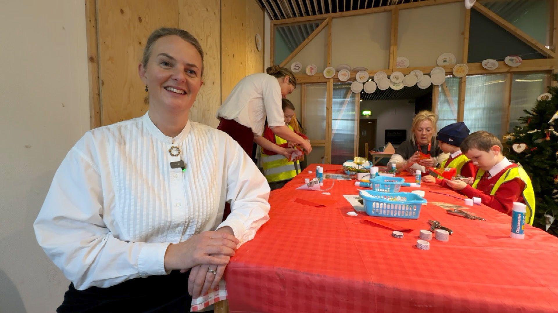 Children wearing high-viz jackets sit at a long table with a red table cloth while crafting, in the foreground a woman smiles as she wears a white Victorian-style shirt and broach. 