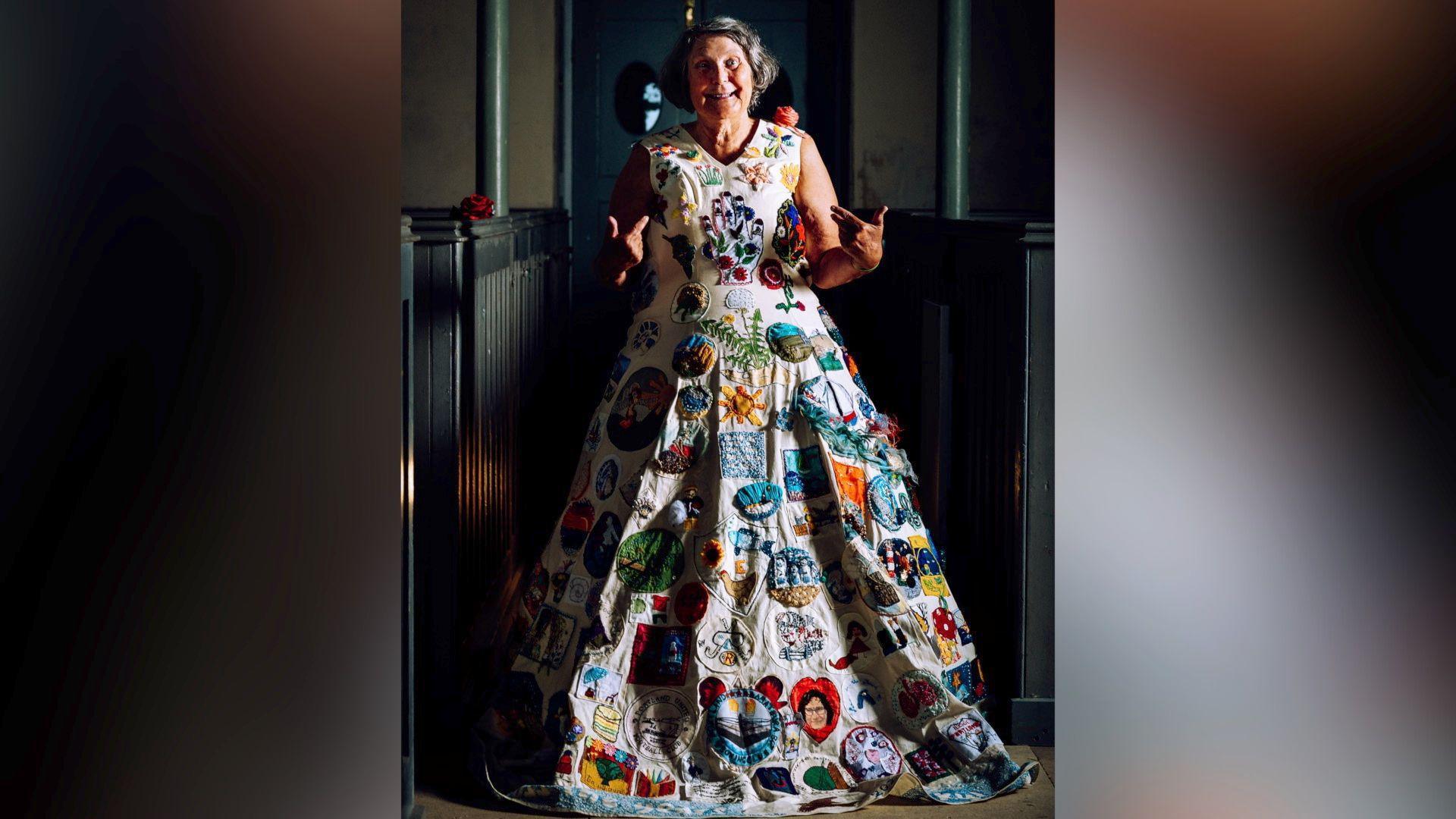 Hazel Smith has grey hair styled in a short bob, she is smiling at the camera and is pointing at herself with both hands. She wears the Portland Dress, which is a white calico dress - a shoulderless v neck top with a full, floorlength skirt, covered in different coloured embroidered patches. She is standing in a corridor, with black wooden stair railings behind her.