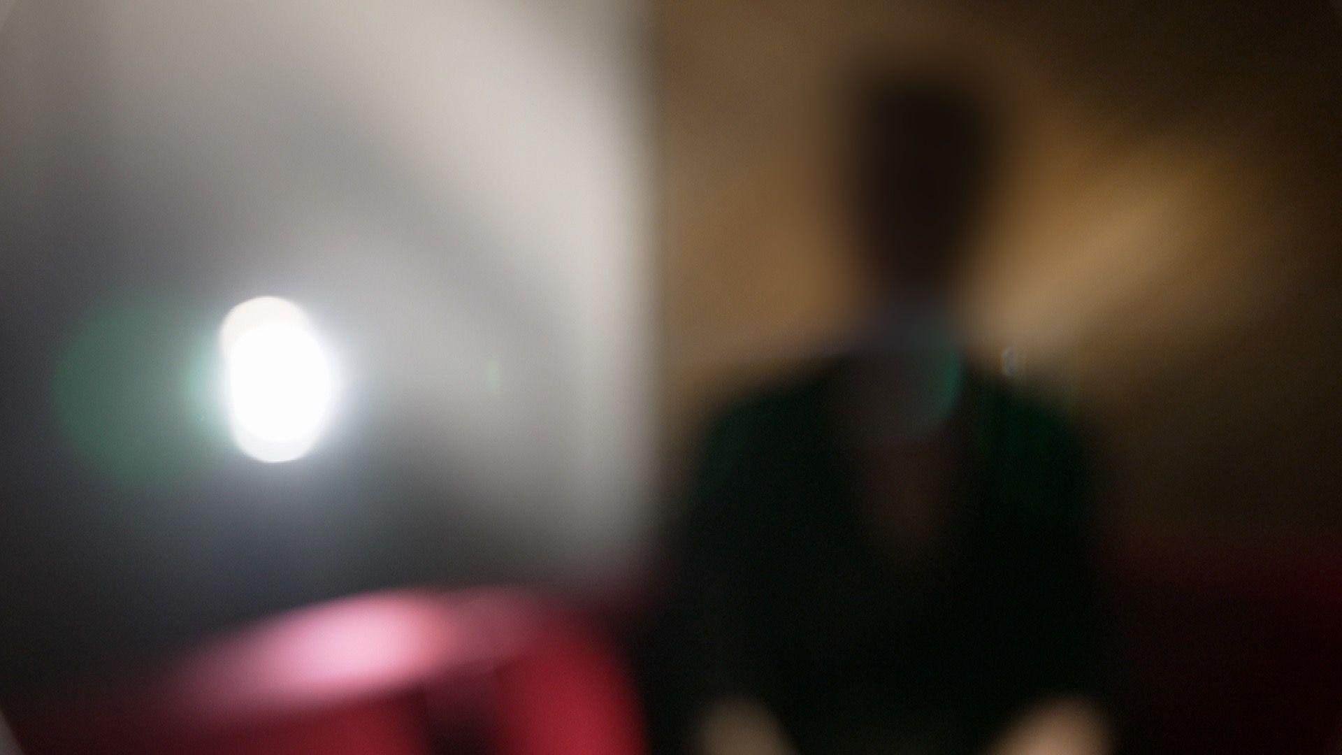 Emma sits indoors, deliberately photographed heavily out of focus so that her face and identifying features cannot be seen. A bright light is visible to the left and a red sofa is behind her.