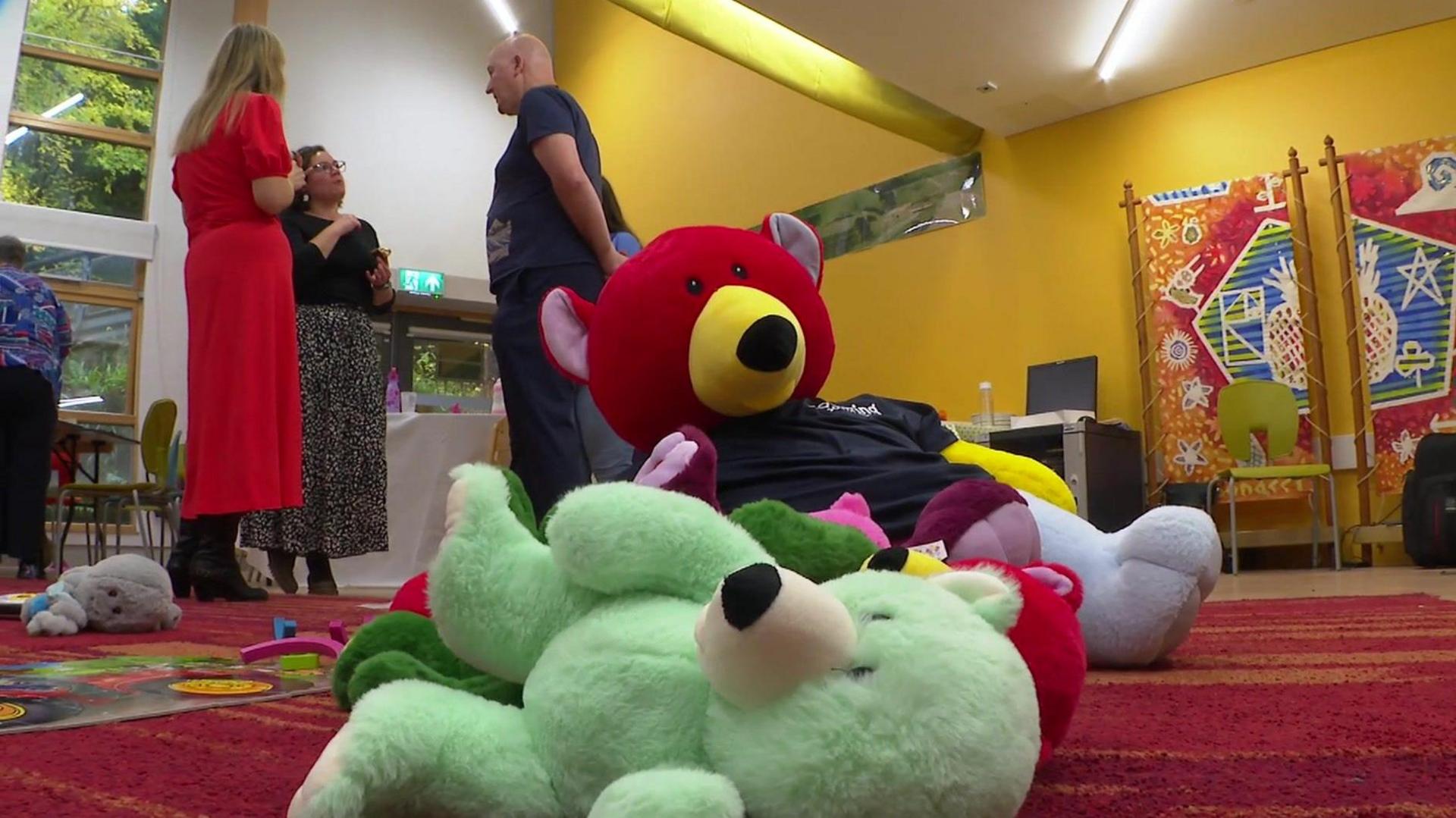 A pile of brightly coloured teddy bears laid on the floor of a children's centre. There are three people in the background talking. 