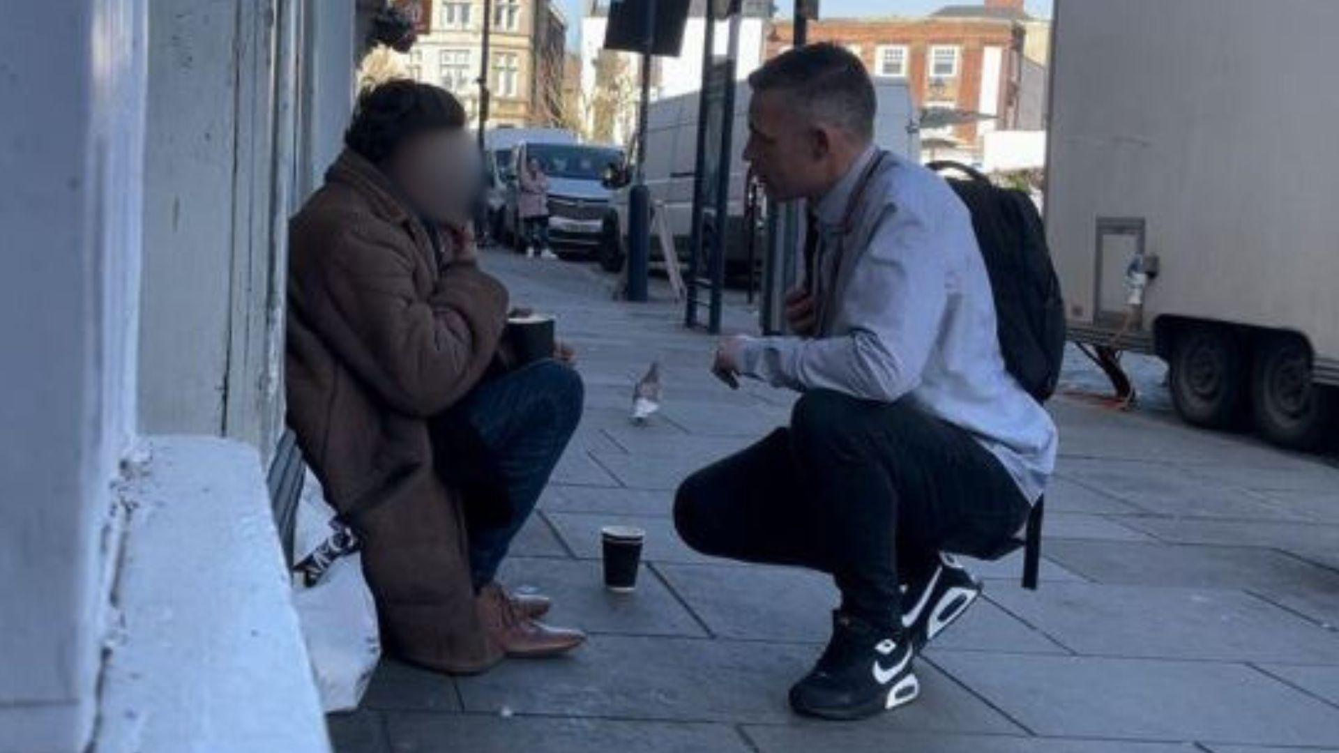 A man wearing a light-coloured shirt with blue jeans and black and white trainers is bending down and speaking to a rough sleeper, who is sitting on a pavement in the street. 