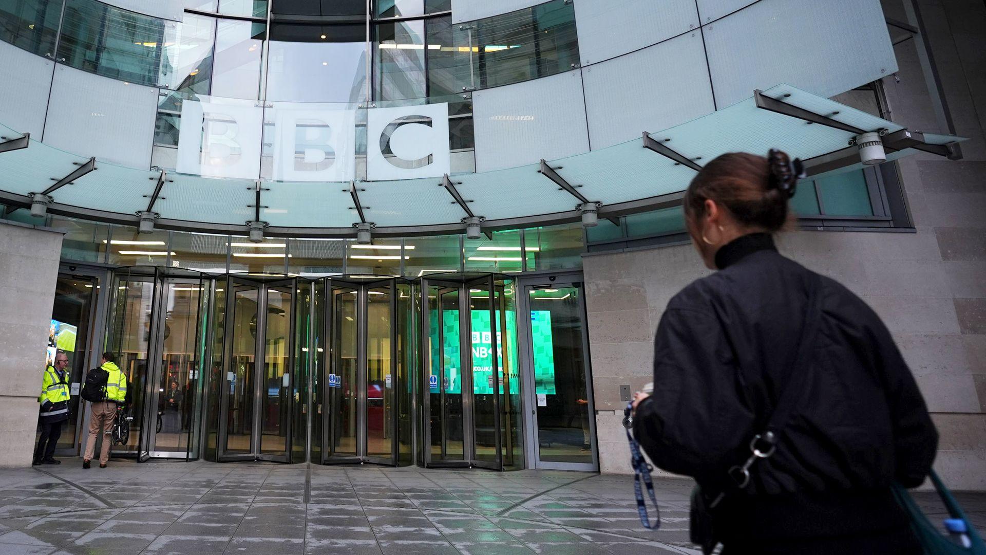 A person wearing a black jacket and carrying a backpack walks towards the entrance doors at BBC Broadcasting House in London on Monday, with the BBC logo in view.