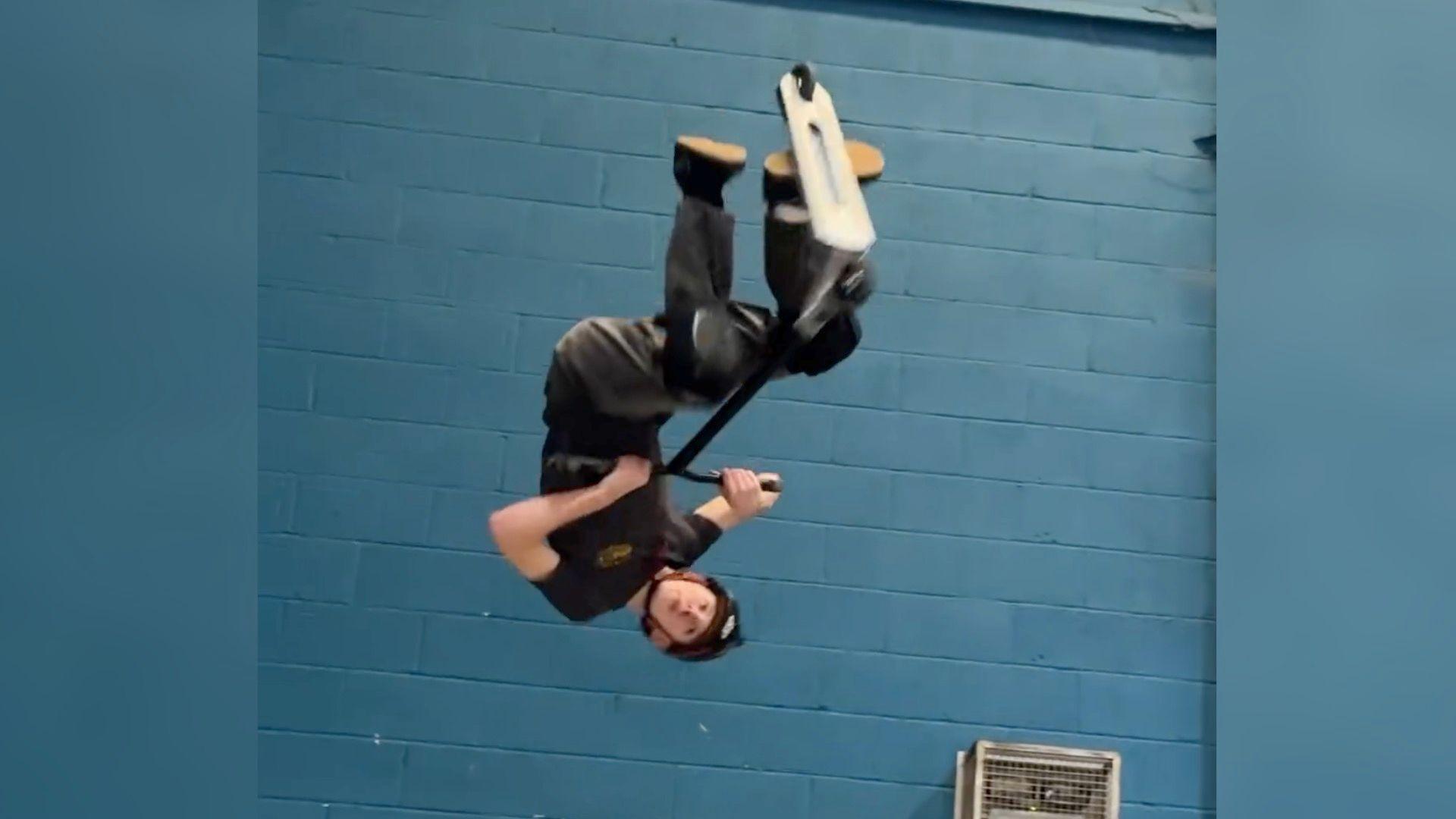 Josh Popham upside down on a scooter in front of a blue wall