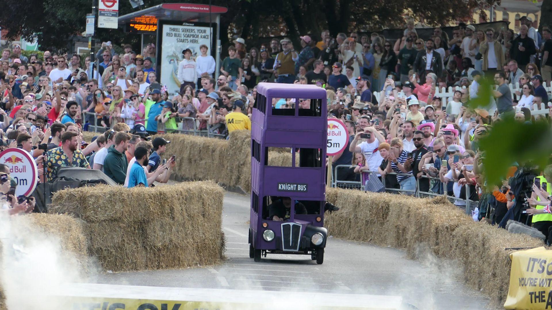 A homemade soapbox race cart, in the style of the Harry Potter knight bus, racing down a track in between hay bales with a crowd watching