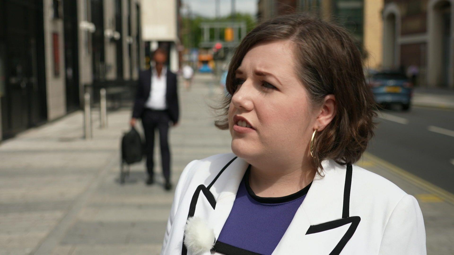 Jade Dyas has short dark hair and is wearing a white jacket with black-trimmed lapels. She is wearing a purple t-shirt underneath. She is standing on Victoria Street in Liverpool.