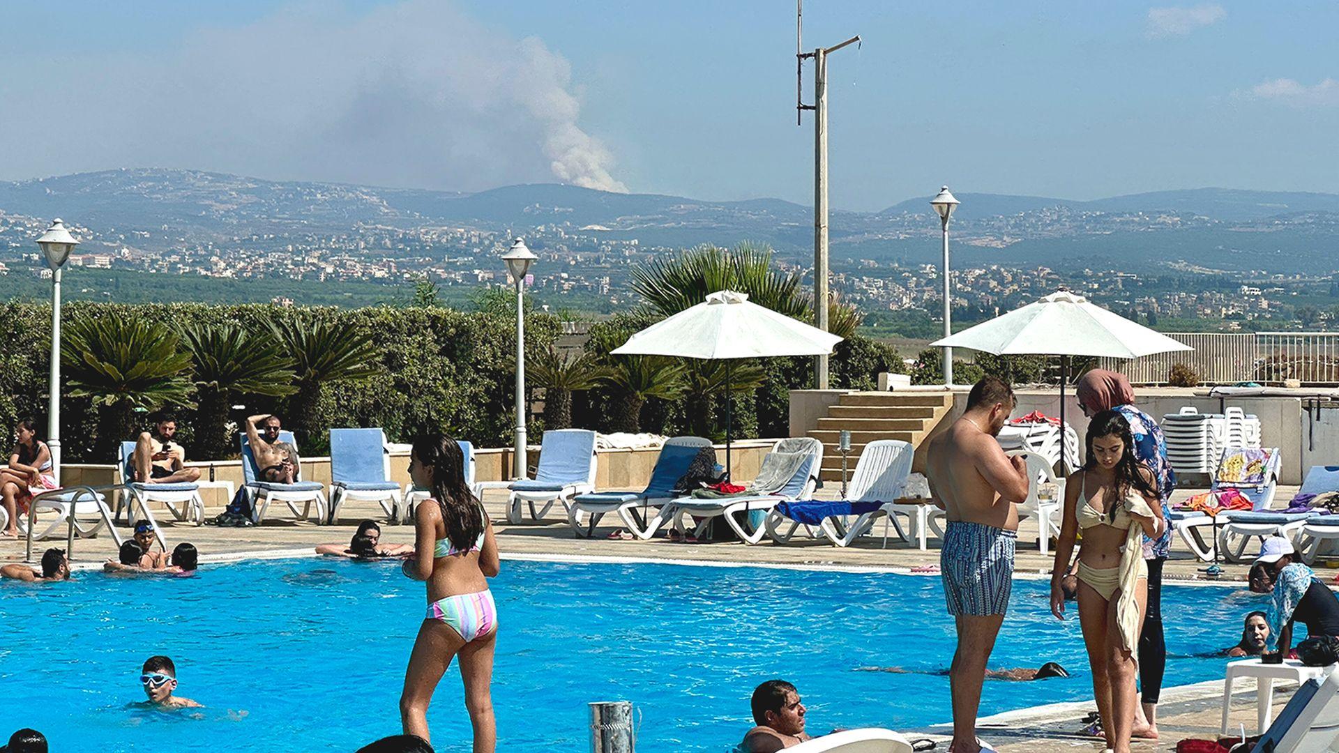 People sunbathe and swim in a resort hotel as explosions smoke billows from a hillside in the distance after an Israeli strike 