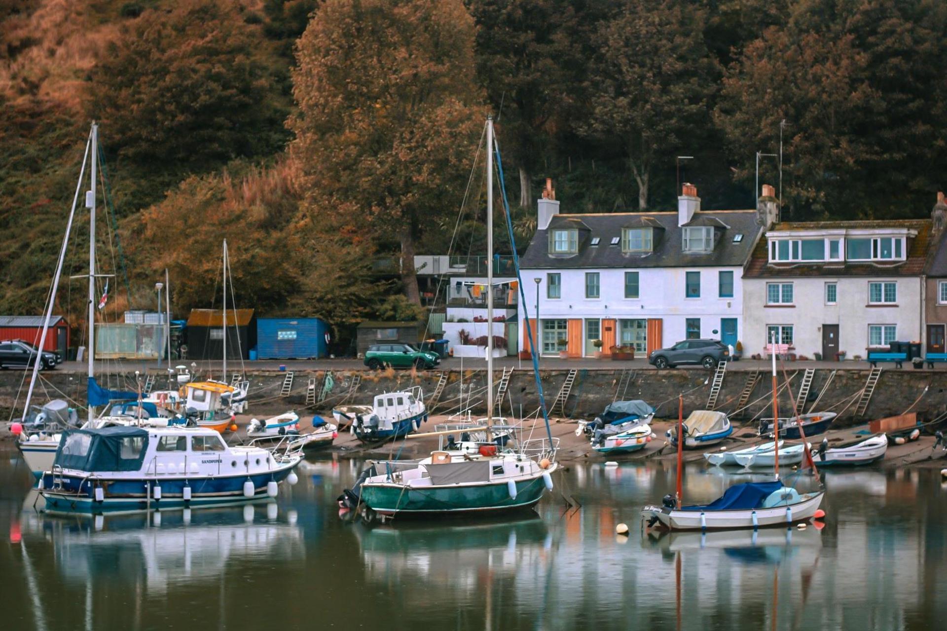 Boats are reflected in the water of the harbour. There are houses and a tree-covered hillside.