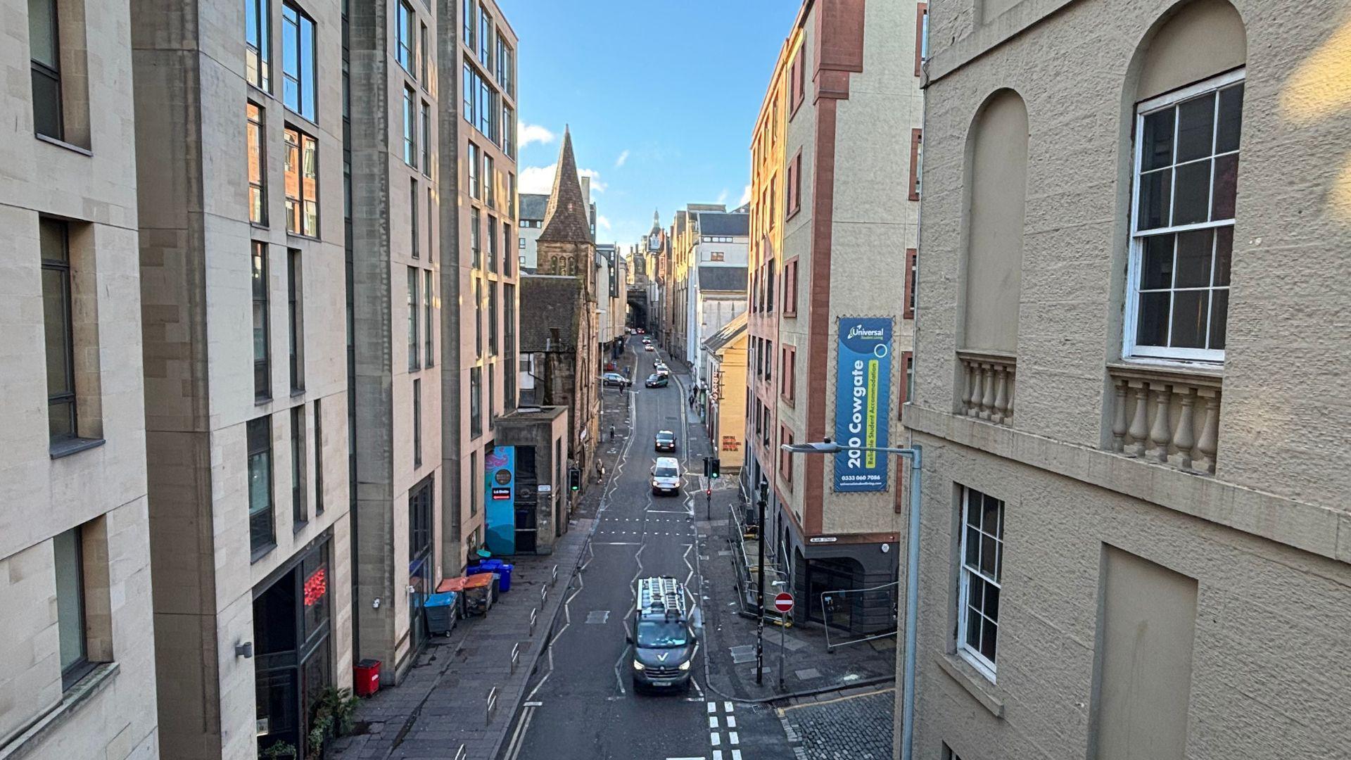 A general view of the Cowgate from South Bridge. Light stone-coloured buildings are on either side with a road running through the middle. A black taxi is in the foreground of the image with several other cars behind it. The sky is blue and it is a sunny day.