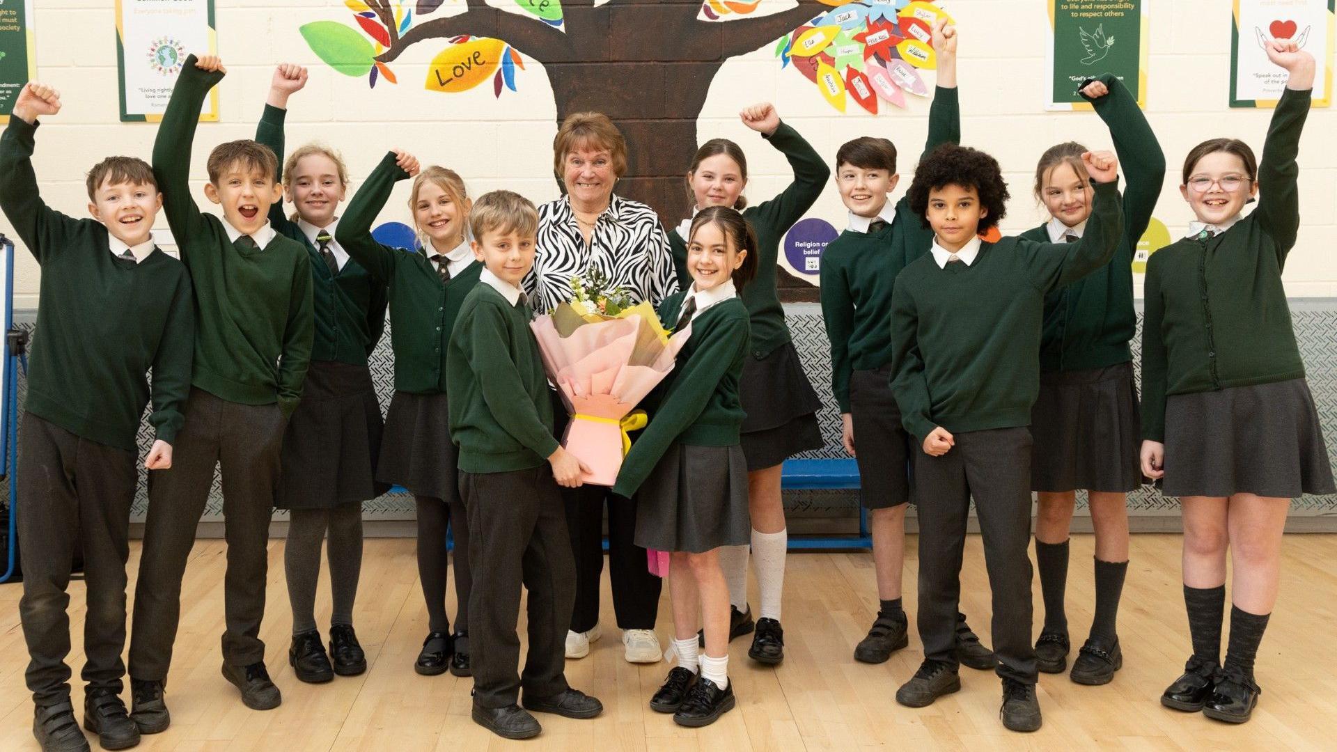 Mrs Hewlett stands in the middle of nine children, two stand in front of her holding a bouquet of flowers. The children wear green jumpers and grey skirts and trousers. Mrs Hewlett wears a zebra print blouse.