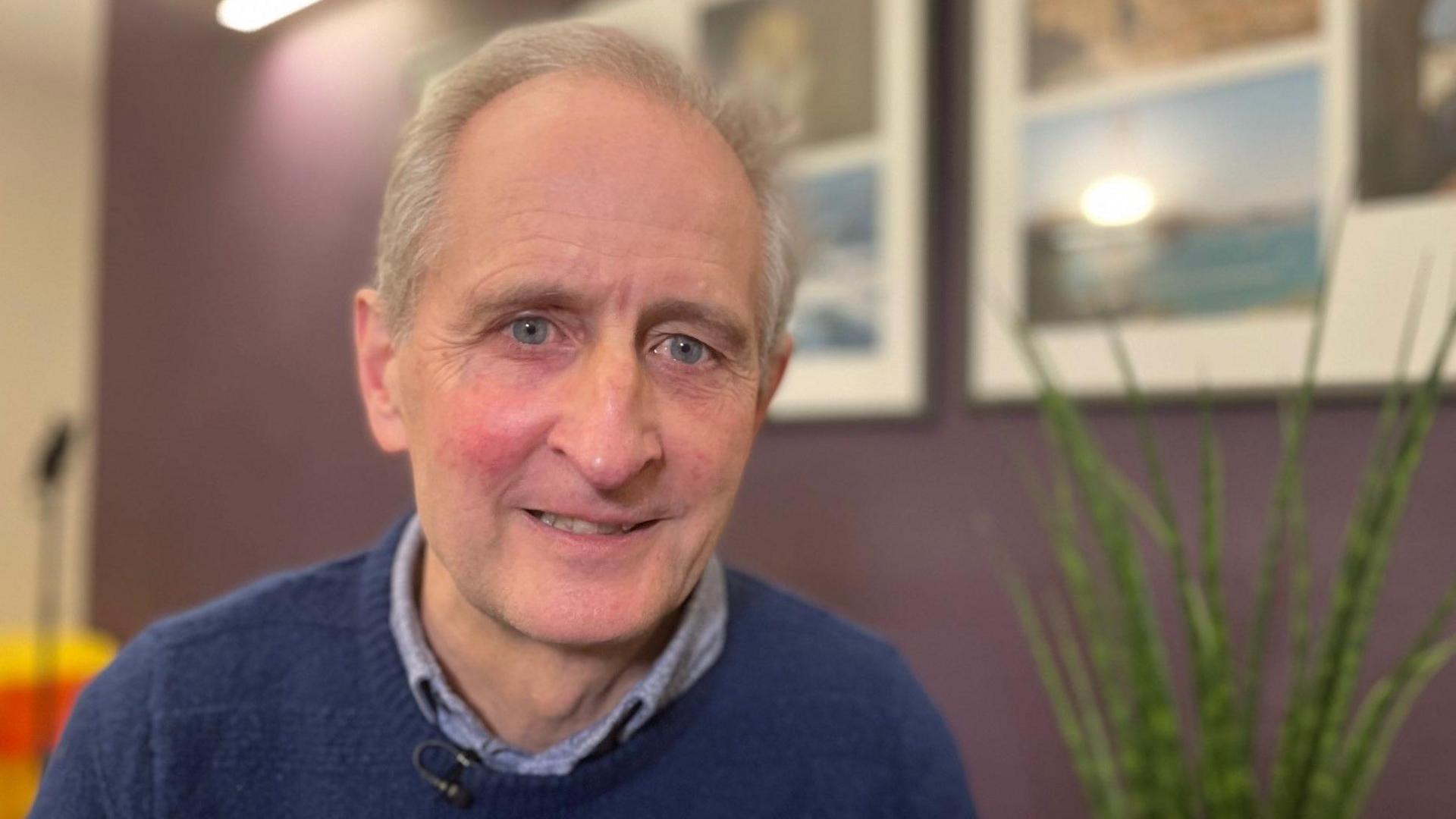 David Steele is looking at the camera. He has grey balding hair and is wearing a blue jumper over a blue shirt. There is a plant behind him and pictures on the wall