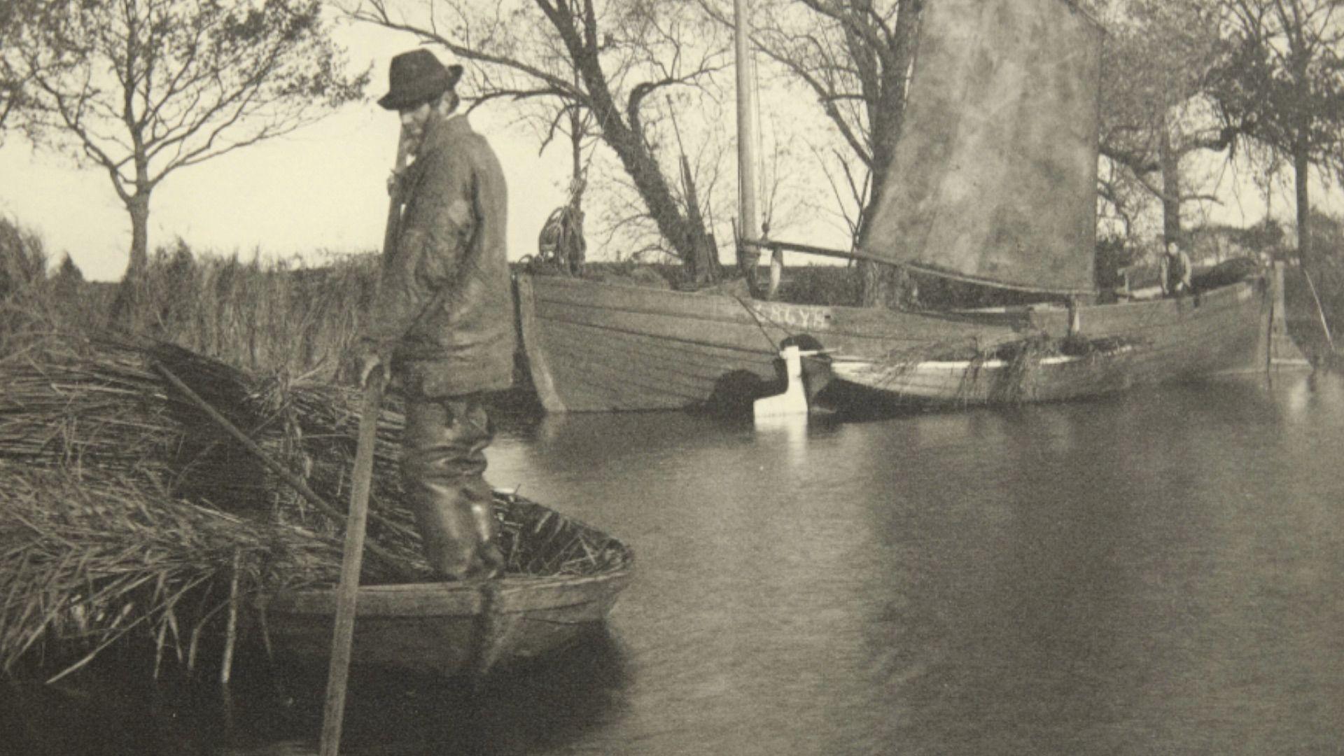 Black and white image of a man pushing through a river on a small boat loaded with reeds with a small wherry boat in the background