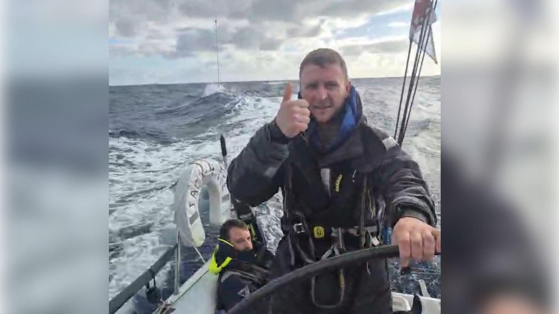 Llun o Thomas Tudor yn codi bawb tra'n llwyio'r cwch ar y mor yn y ras hwylio ar draws yr Iwerydd