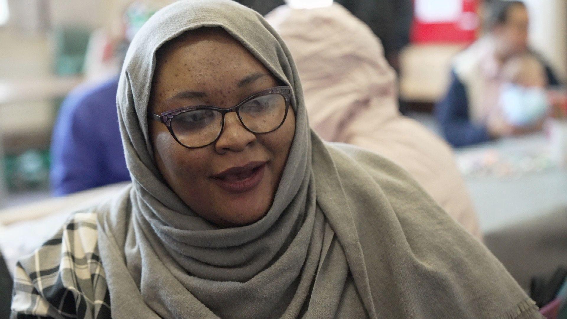 A woman wearing horn-rimmed glasses and a grey headscarf sits in front of other asylum seekers, including a mother and baby.