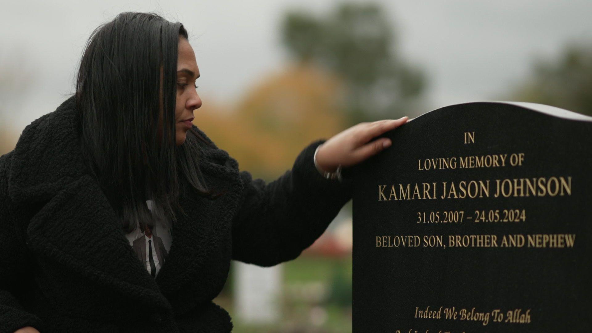 Katherine Johnson, wearing a black coat and a T-shirt with a photo of her son on it, kneels by his gravestone, which bears his name and refers to him as a 'beloved son, brother and nephew' 
