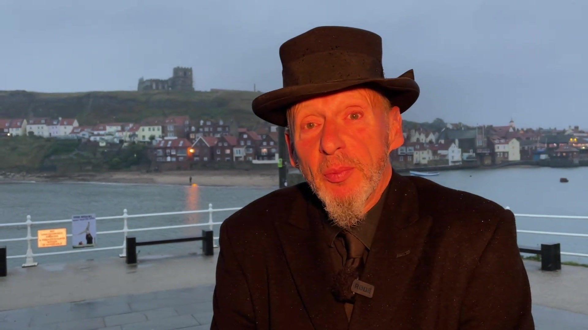 A man in a black suit and hat standing on the seafront at Whitby, behind him Whitby Abbey can be seen on top of the hill.