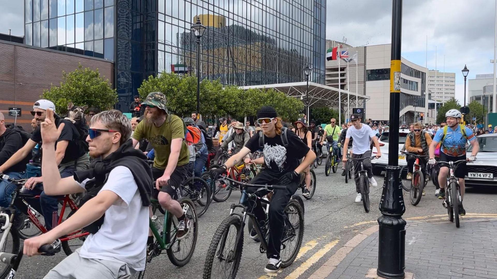 A group of men are cycling on their bikes, some are wearing helmets and glasses. They are dancing along to music as they cycle on a road.