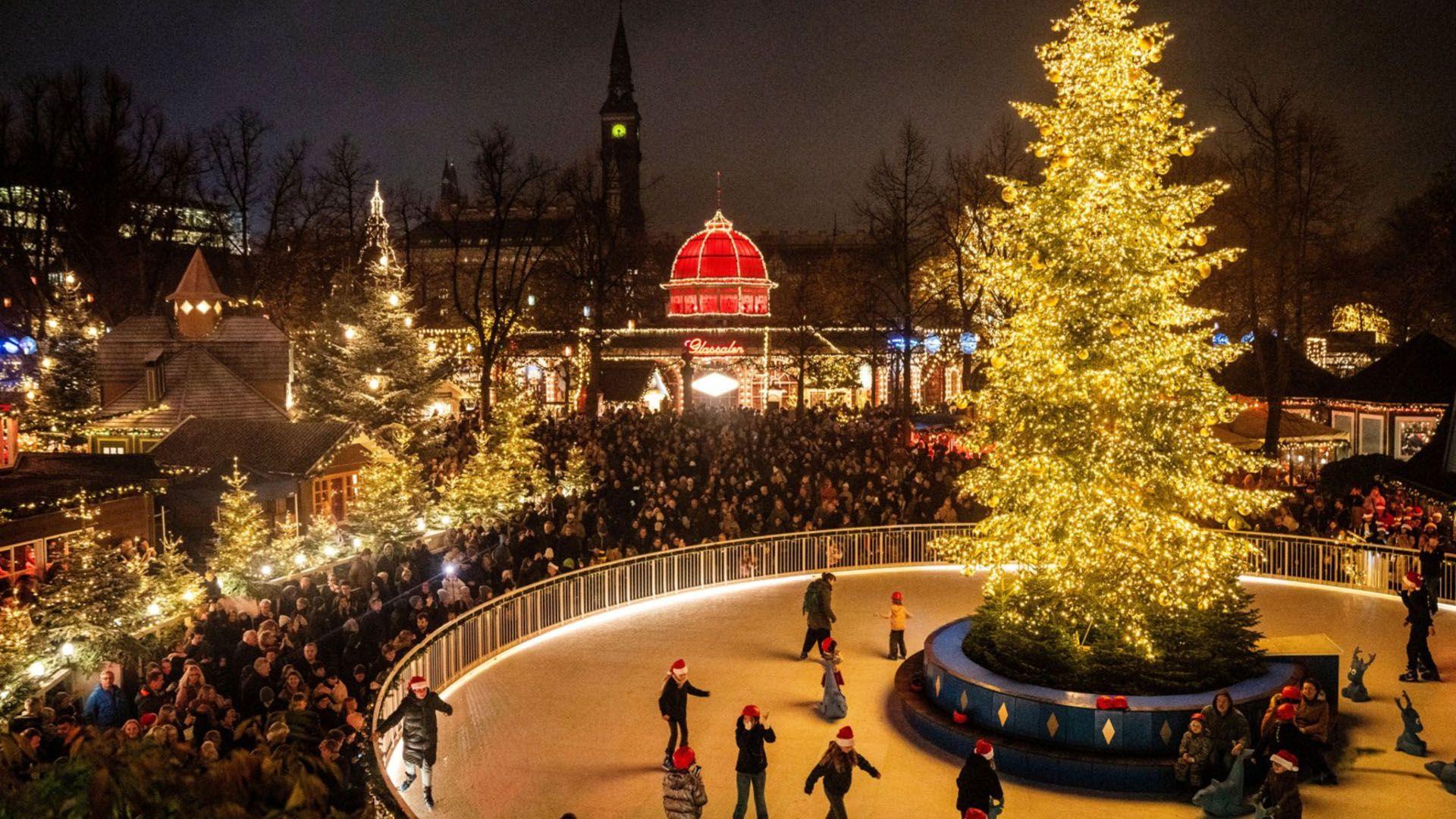 People ice skate in a circular ice rink with a christmas tree in the centre - crowds look on watching the ice skaters from outside the rink