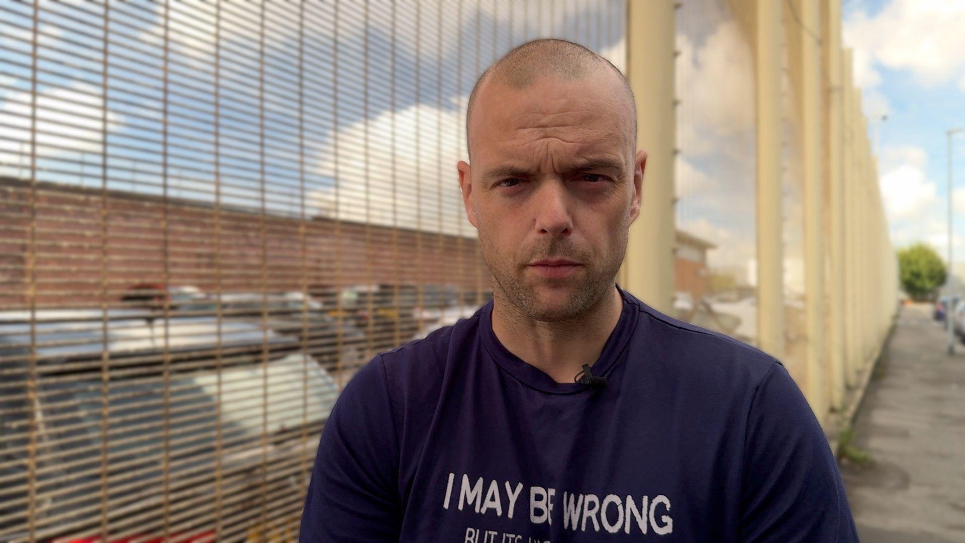 Jamie is wearing a black t-shirt and is standing next to a tall fence. He has a shaved head and stubble. 