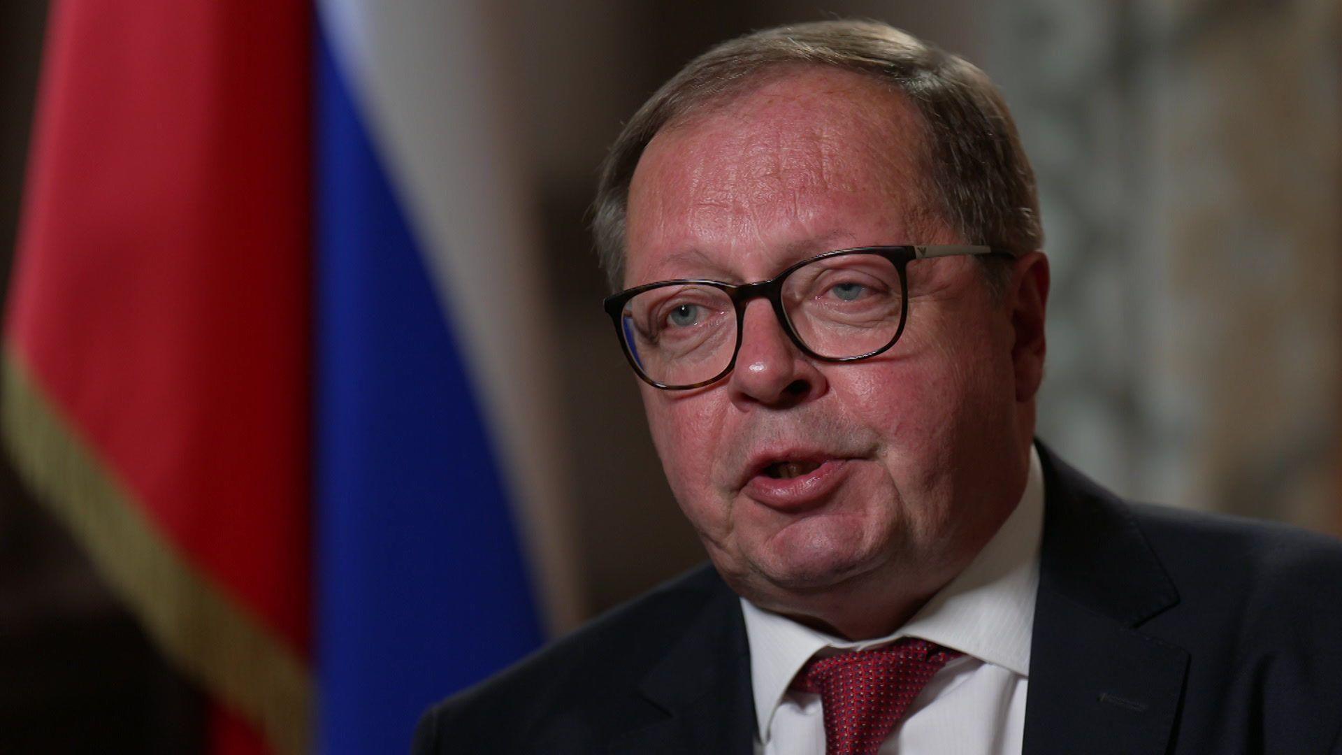 Headshot of Andrei Kelin speaking during an interview with the Russian flag positioned behind him