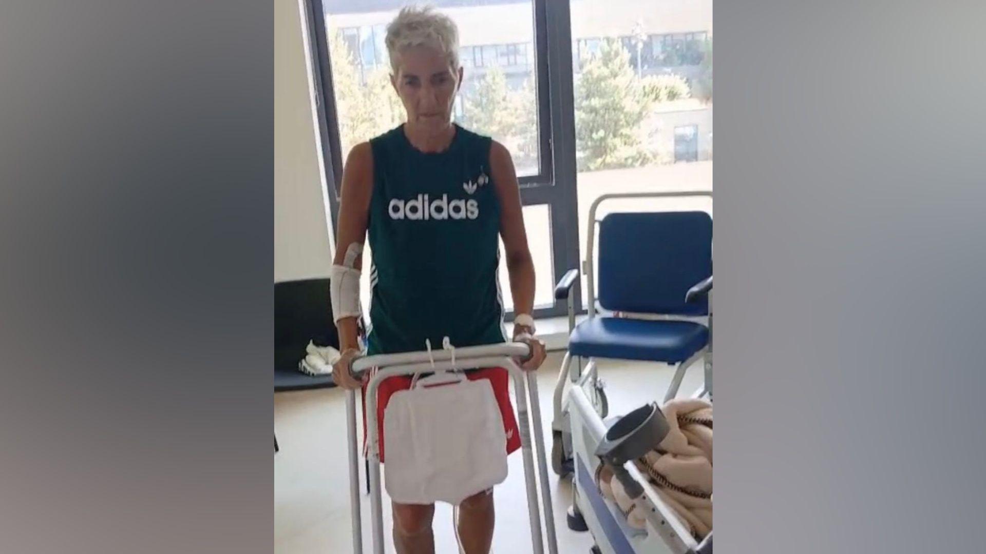 A woman using a zimmer frame to walk. She is wearing a navy vest top and has short grey hair.