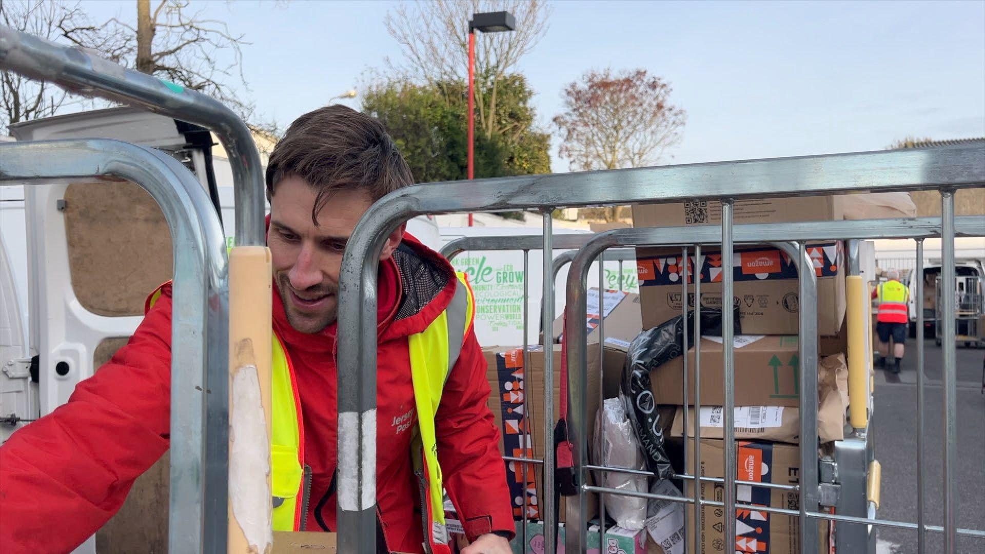 A man with brown hair in a red postal jacket and a high-viz vest reaching for a package. He is behind a trolley full of parcels, next to a white postal van.