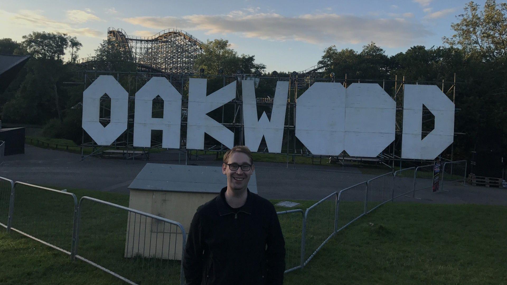 Nick Hutson standing in front of a large set of white letters spelling Oakwood at the theme park. In the background you can see the Megafobia ride above the treeline. Nick is wearing a black jacket and has blond short hair and glasses.