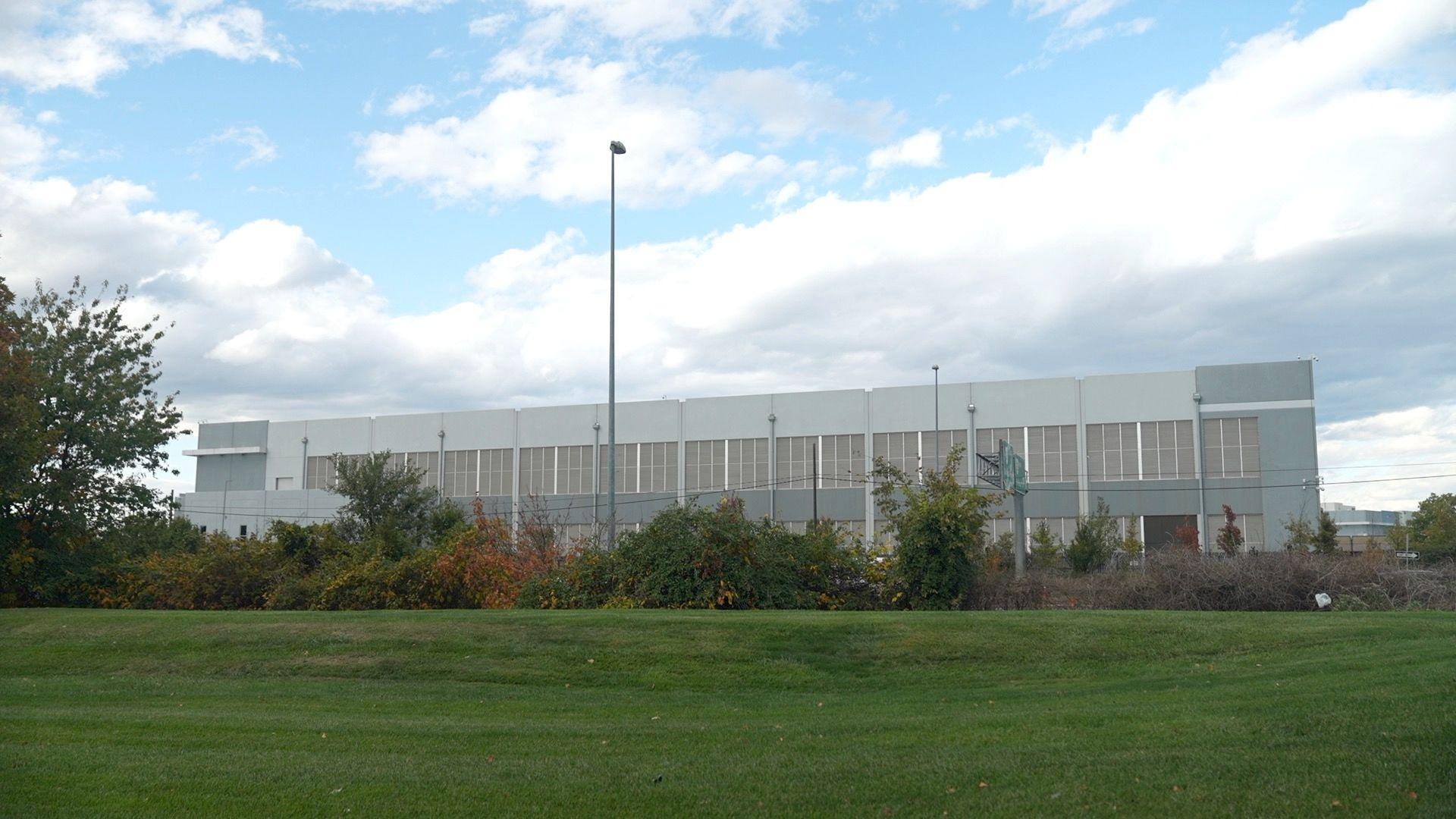 A large industrial warehouse-type building sits behind a flat grassy field and trees.