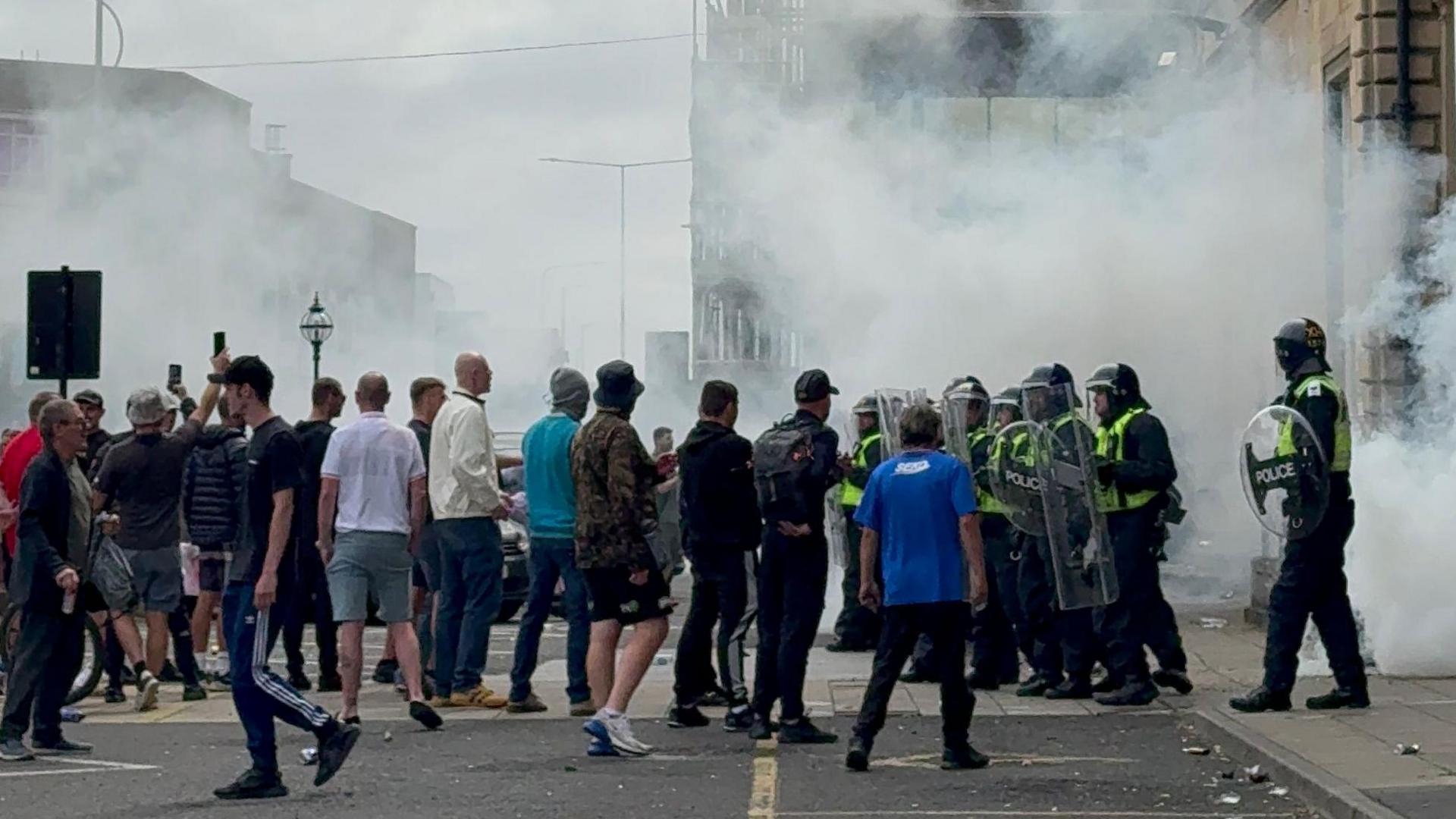 A crowd of people walking towards police officers wearing riot gear