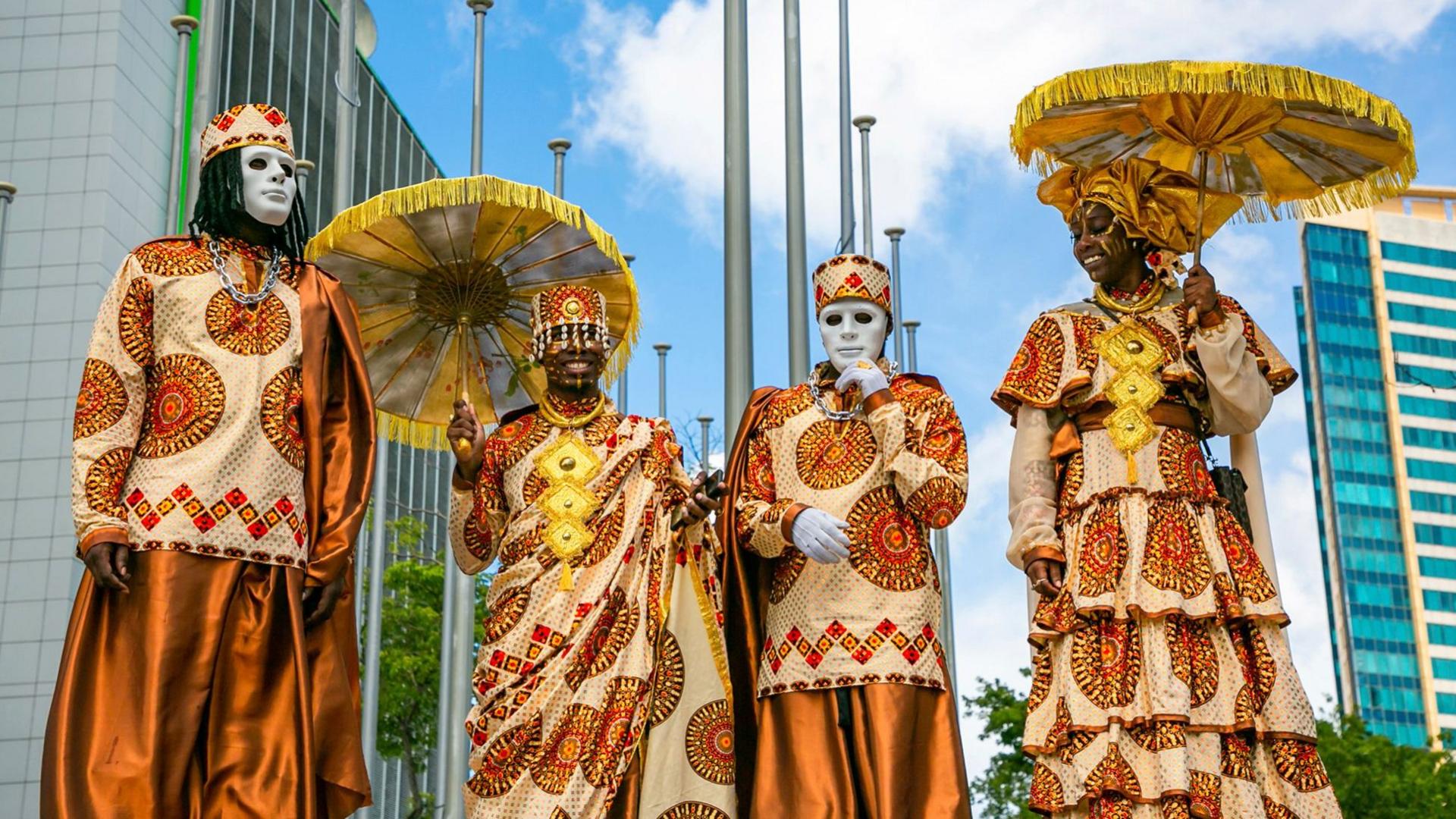 Four people stand in a row, wearing African print outfits. Two wear white masks, the other two carry parasol