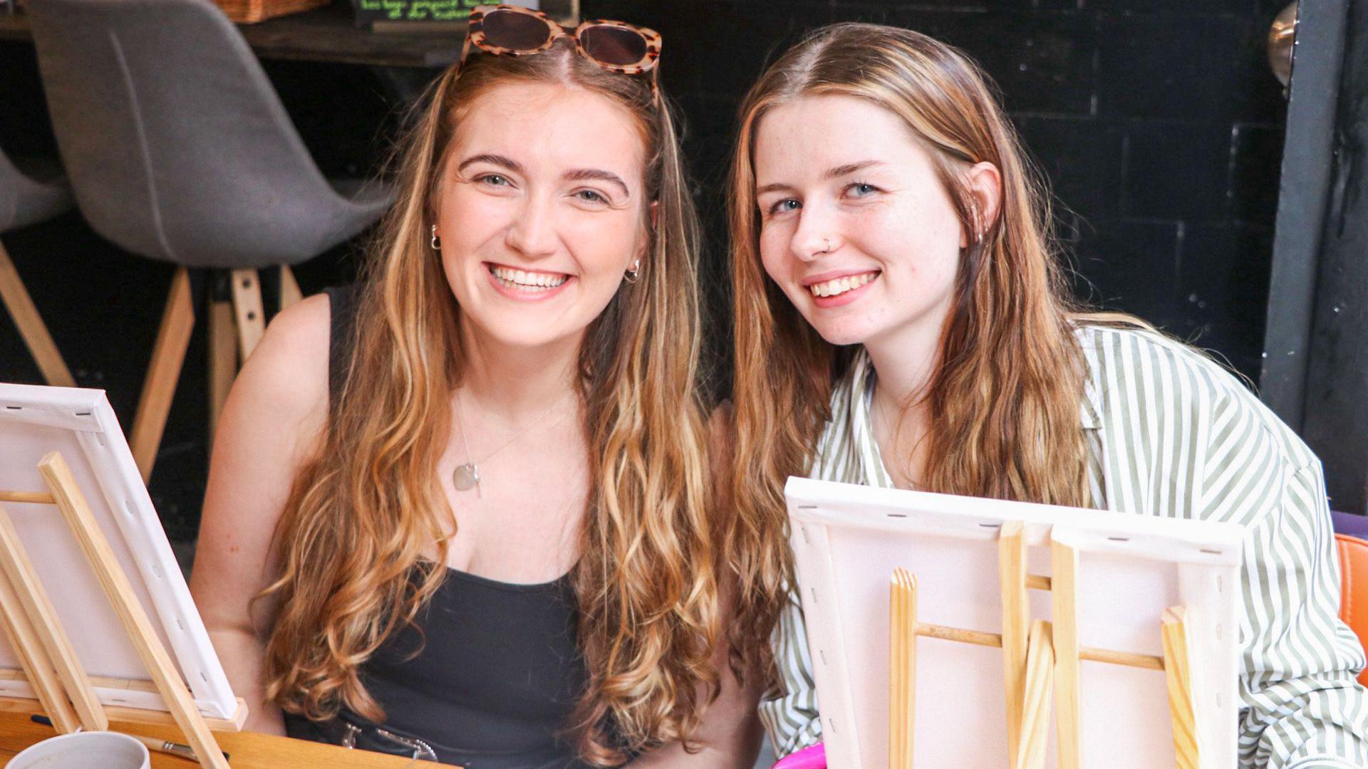 Tiffany Baker Quayle and Caitlin Ball smiling while sitting in front of two small easels. Tiffany is wearing sunglasses and a black tank-top, while Caitlin is wearing a green and white striped shirt. Both have long light brown hair.