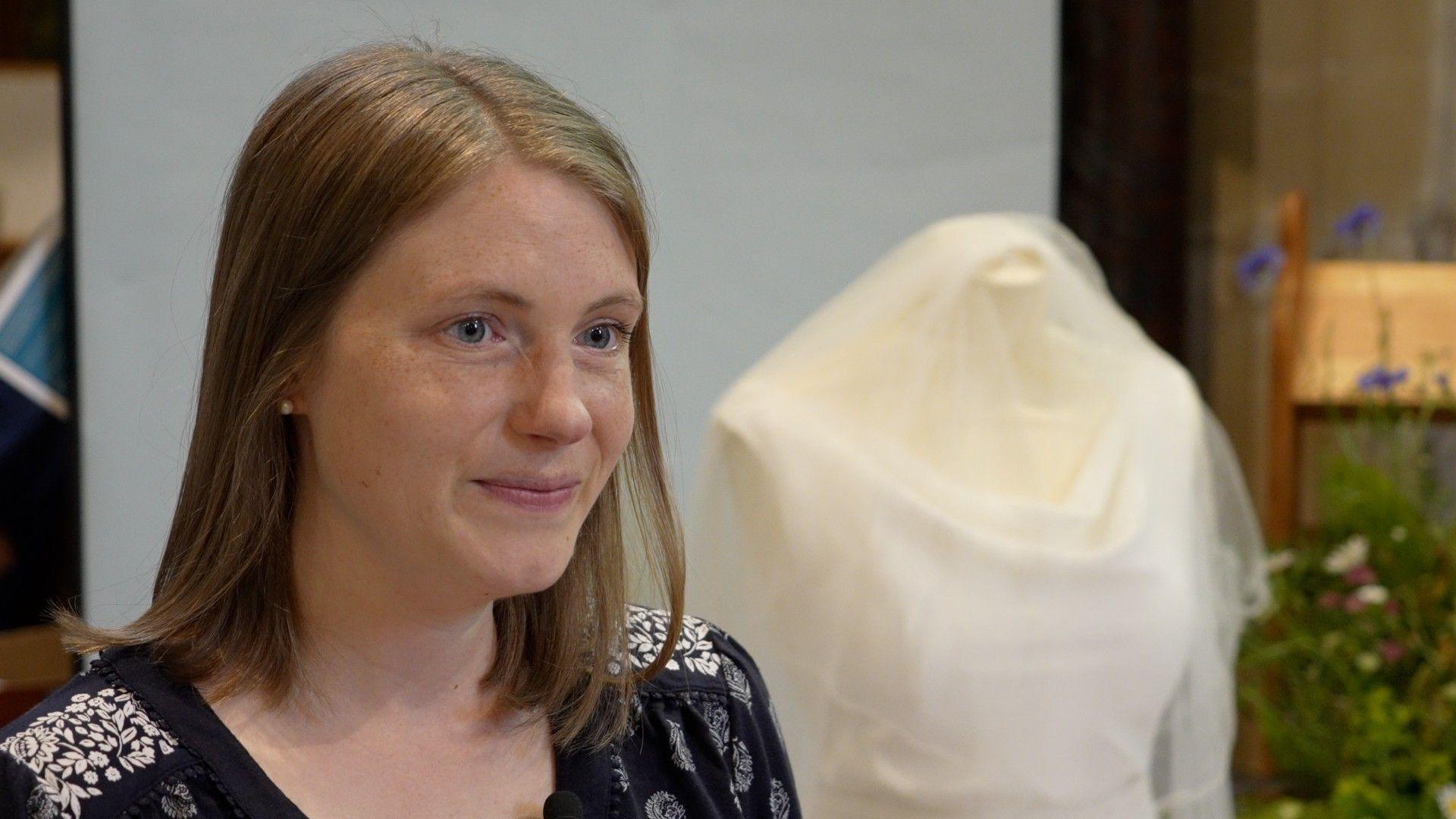 A young woman smiles with a wedding dress in the background.