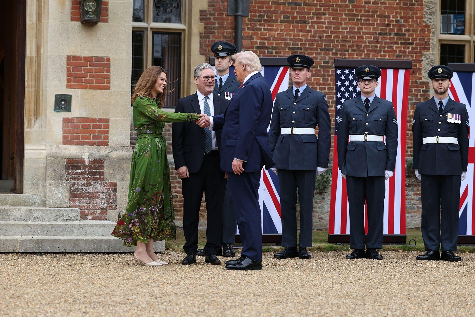 Victoria Starmer (L), wearing a mid-calf length floral green dress, greets US President Donald J. Tump (R) as British Prime Minister Keir Starmer (C) looks on ahead of their talks at Chequers. Four servicemen in uniform can be seen behind along with the US and Union Jack flags.