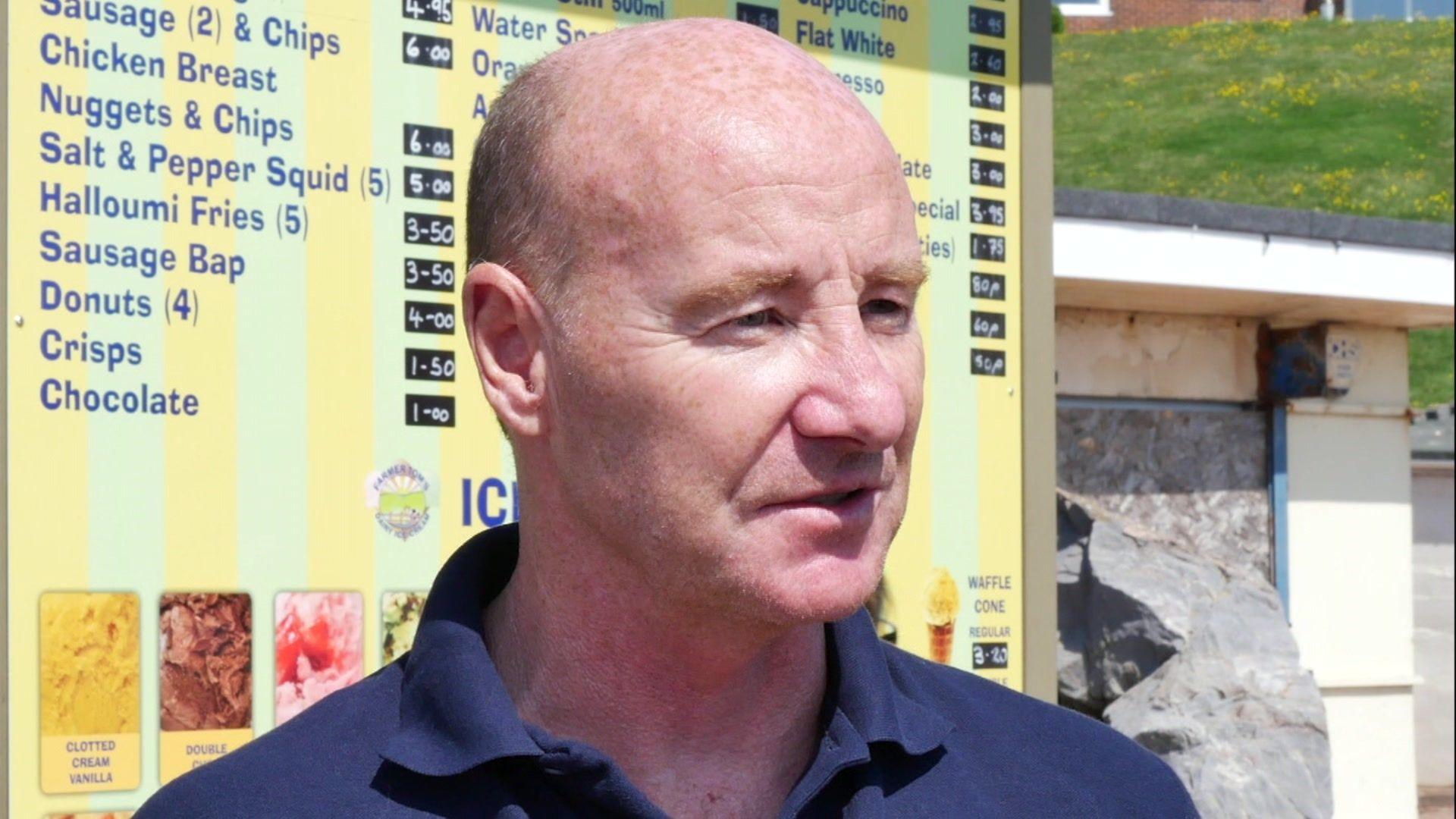 A photo which shows Dave Bamforth stood in front of a menu which lists items such as chicken nuggets and chips and ice cream. He is wearing a blue polo shirt.