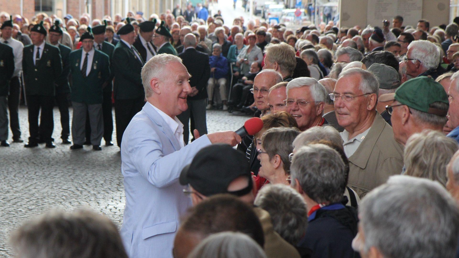 Dave Monk wears a light blue jacket and white shirt. He holds a microphone with a red cover and he is speaking to a crowd of people. Behind Dave is a group of men in uniform who are about to play the Last Post at the Menin Gate in Ypres, Belgium