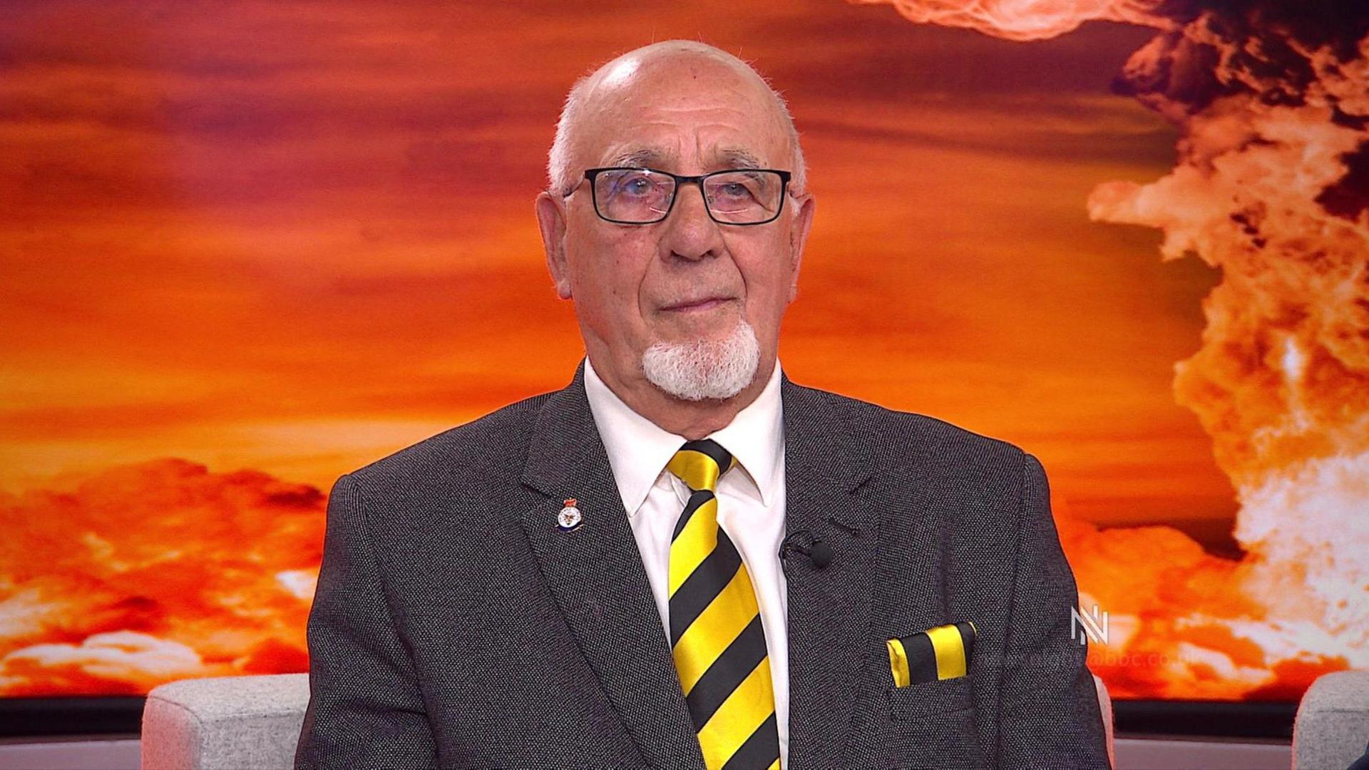 A man sits in a studio with a backdrop of a fiery nuclear test. He is wearing glasses and a white beard and a yellow and black striped tie
