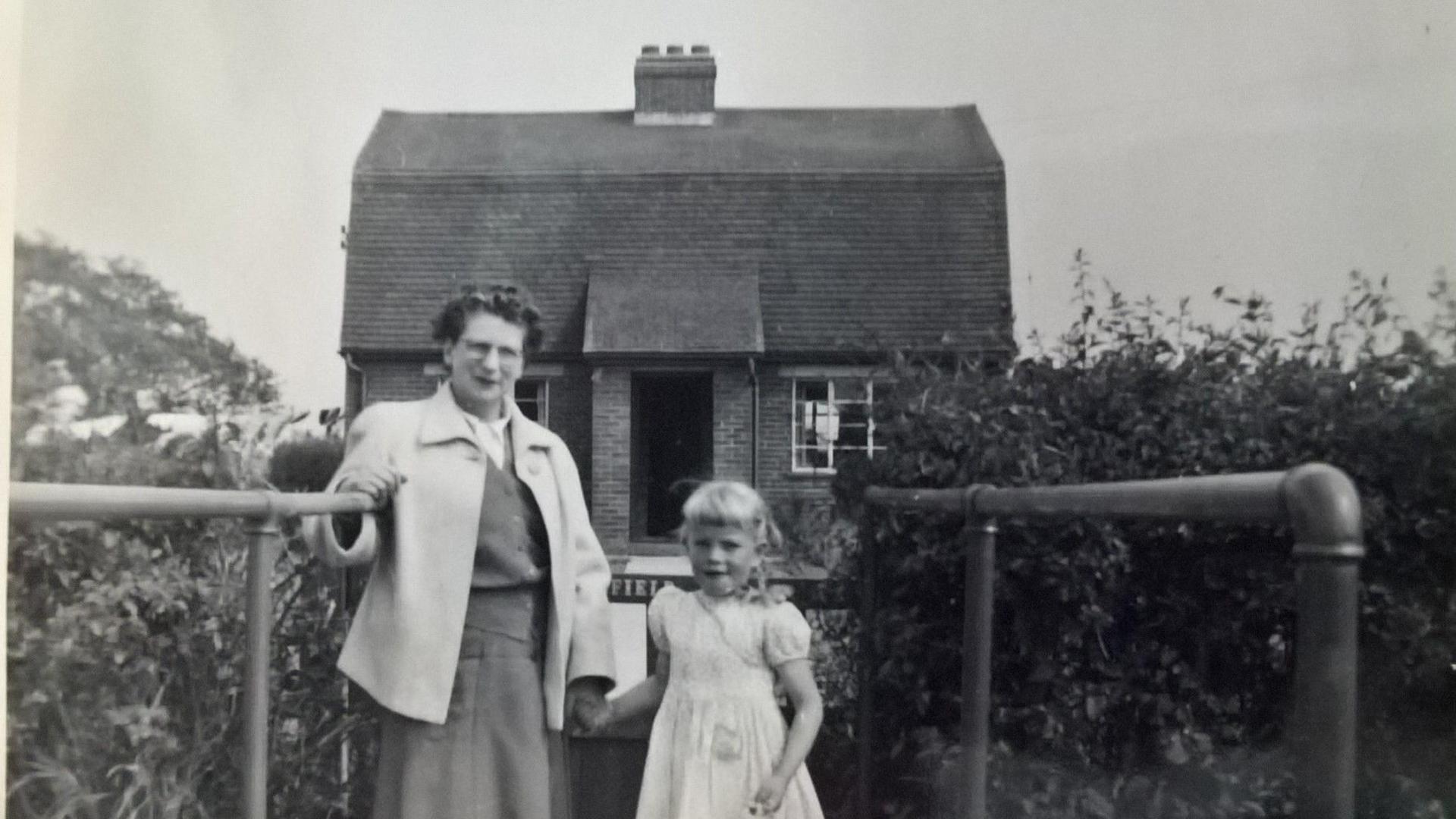 An old black and white photograph of a middle-aged woman and a young girl standing in front of a two-up two-down cottage in the countryside.