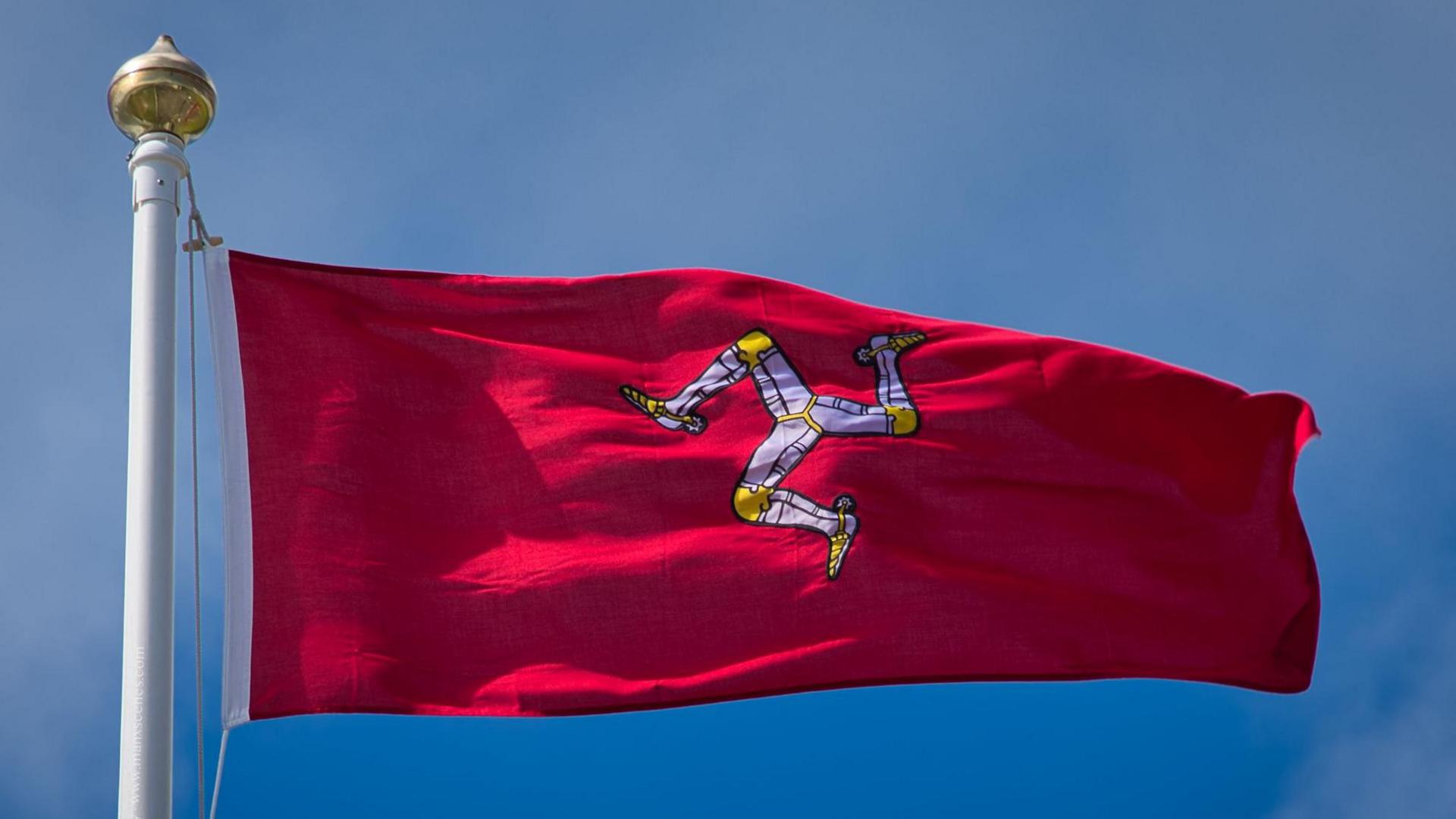 An Isle of Man Flag flying on a flagpole against a blue sky. The flag is red, with a symbol of three armoured legs in the centre. 