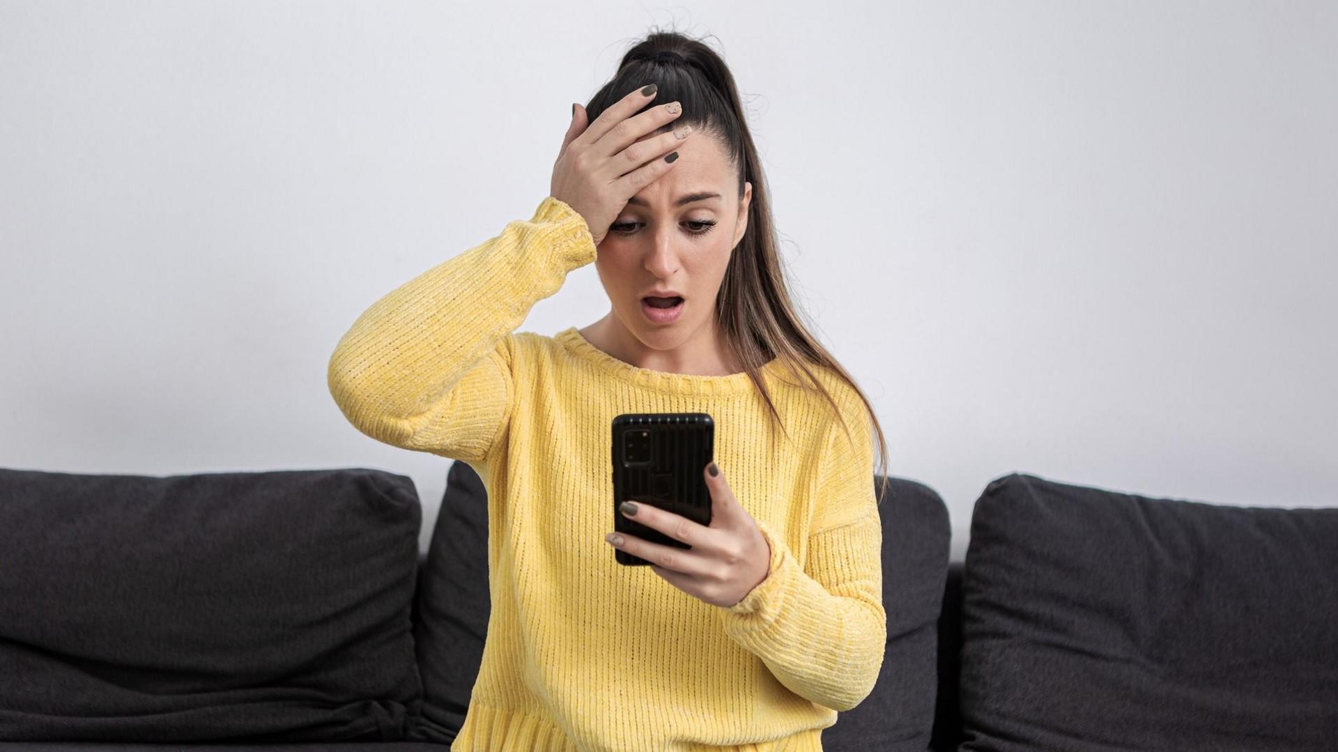 A woman in a yellow jersey, holding a phone, looks shocked 