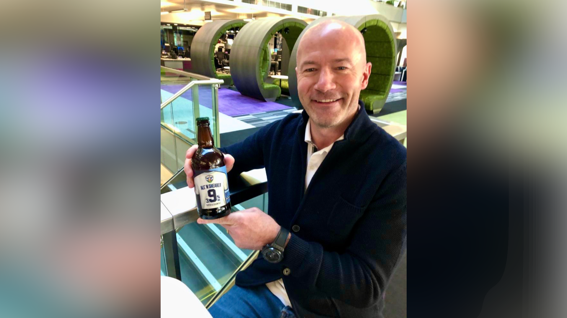 Alan Shearer is pictured holding a bottle of Away Days beer. He is smiling at the camera. He is bald and wearing a navy cardigan with a shirt on underneath. 