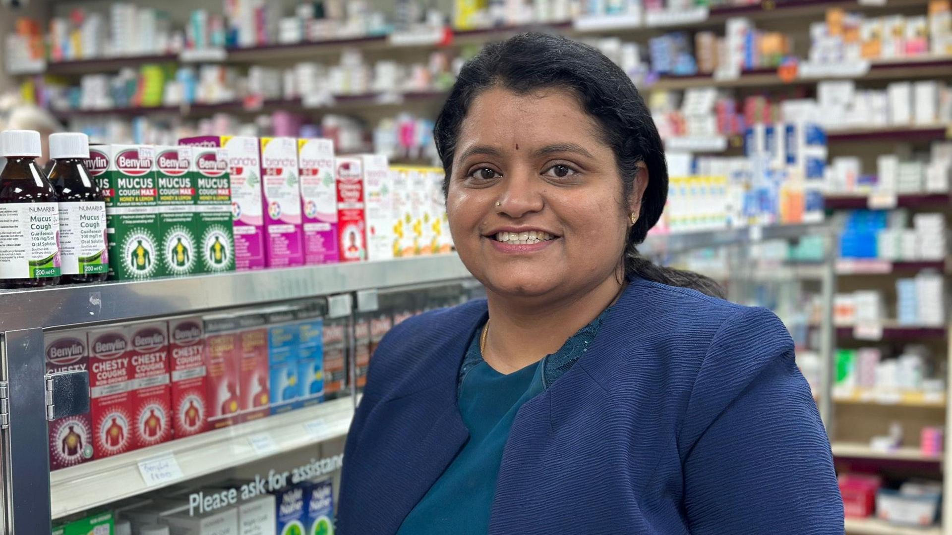 Sri is pictured in her pharmacy in Barmston, standing in front of shelves full of cold and flu medicine. She has long black hair which is tied back from her face. She is wearing a smart blue jacket. 