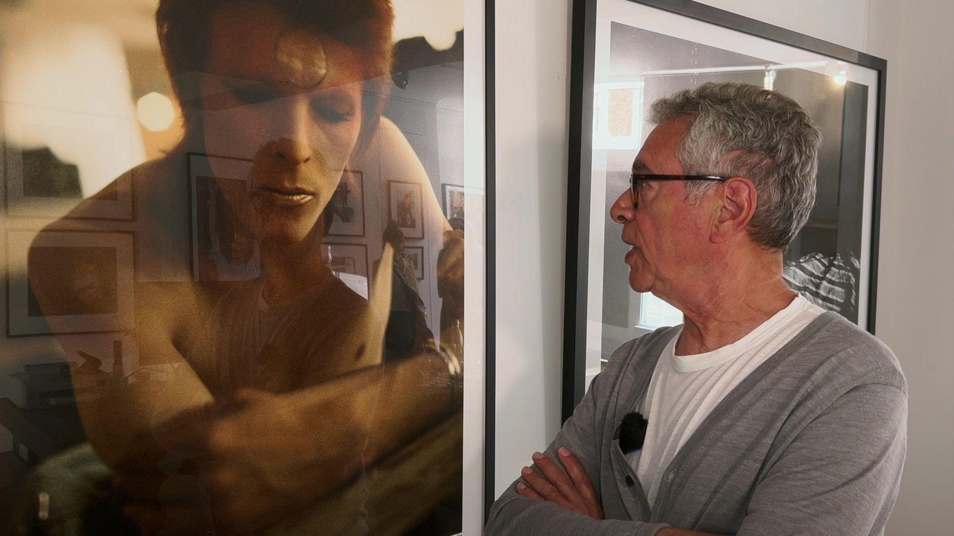 Geoff MacCormack a grey haired man with a grey cardigan and white t-shirt on looking at framed photo of David Bowie on the wall of a gallery 